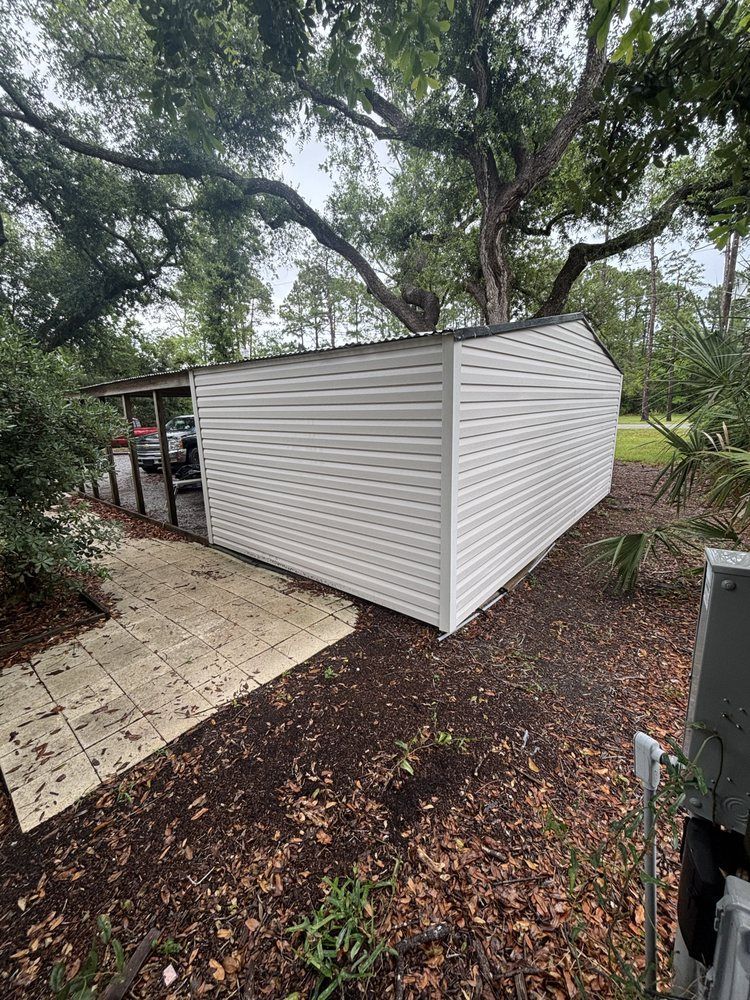 White vinyl shed beside a patio under large trees, with a stone path and mulch around it