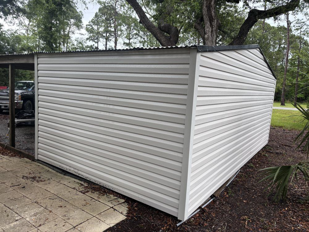 White corrugated metal shed with slanted roof beside a paved area and trees