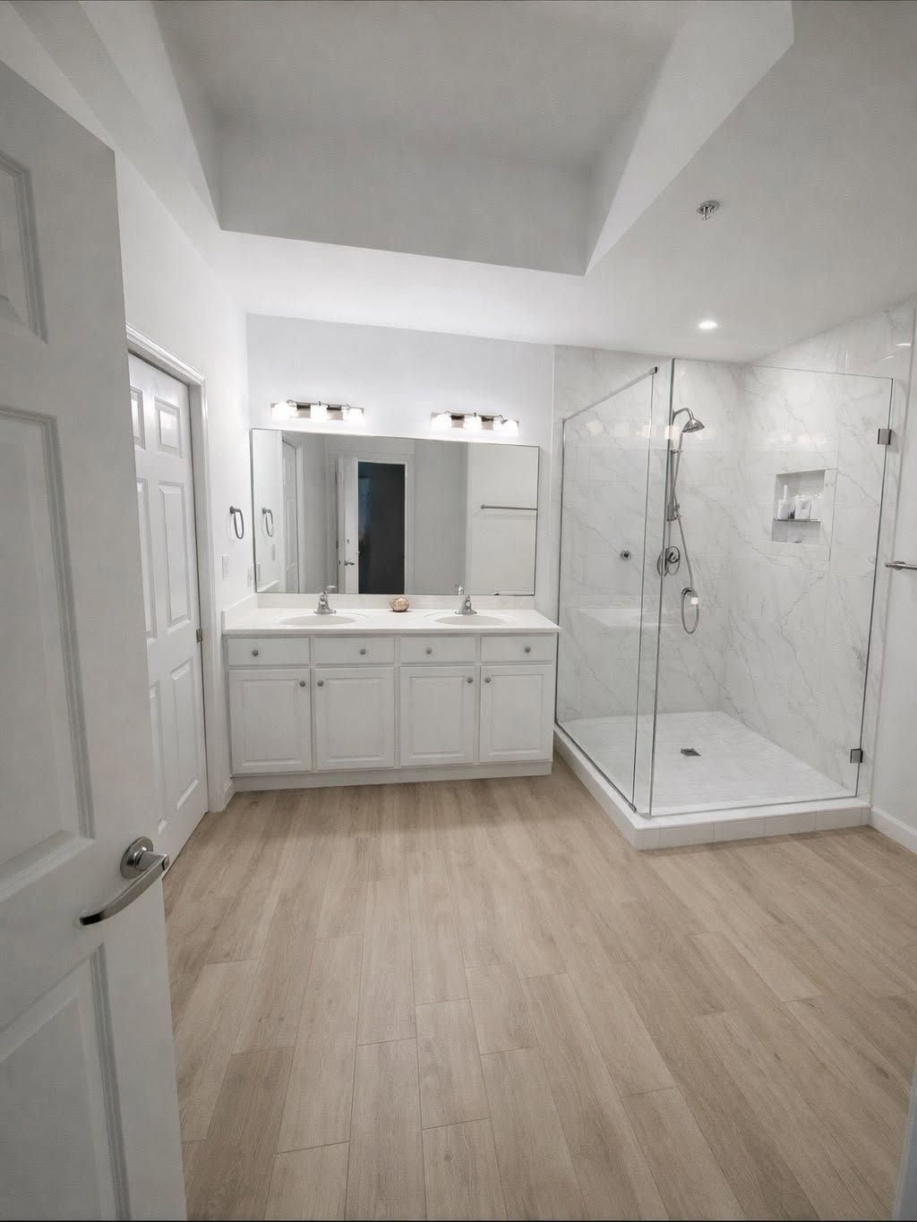 Modern white bathroom with double vanity, large mirror, glass shower, and wood-look floor.