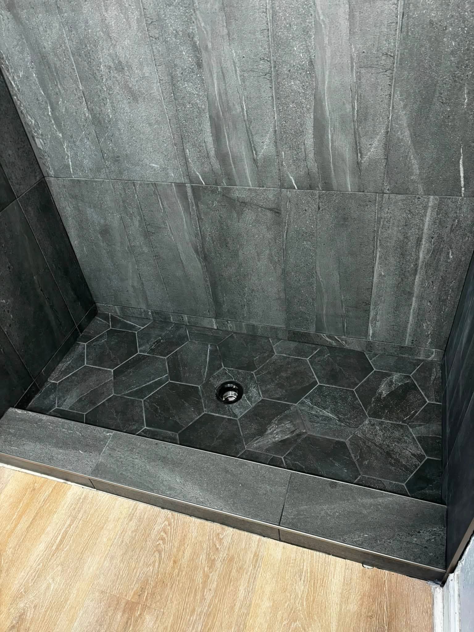 Dark tiled shower stall with a central drain and wood-look floor outside