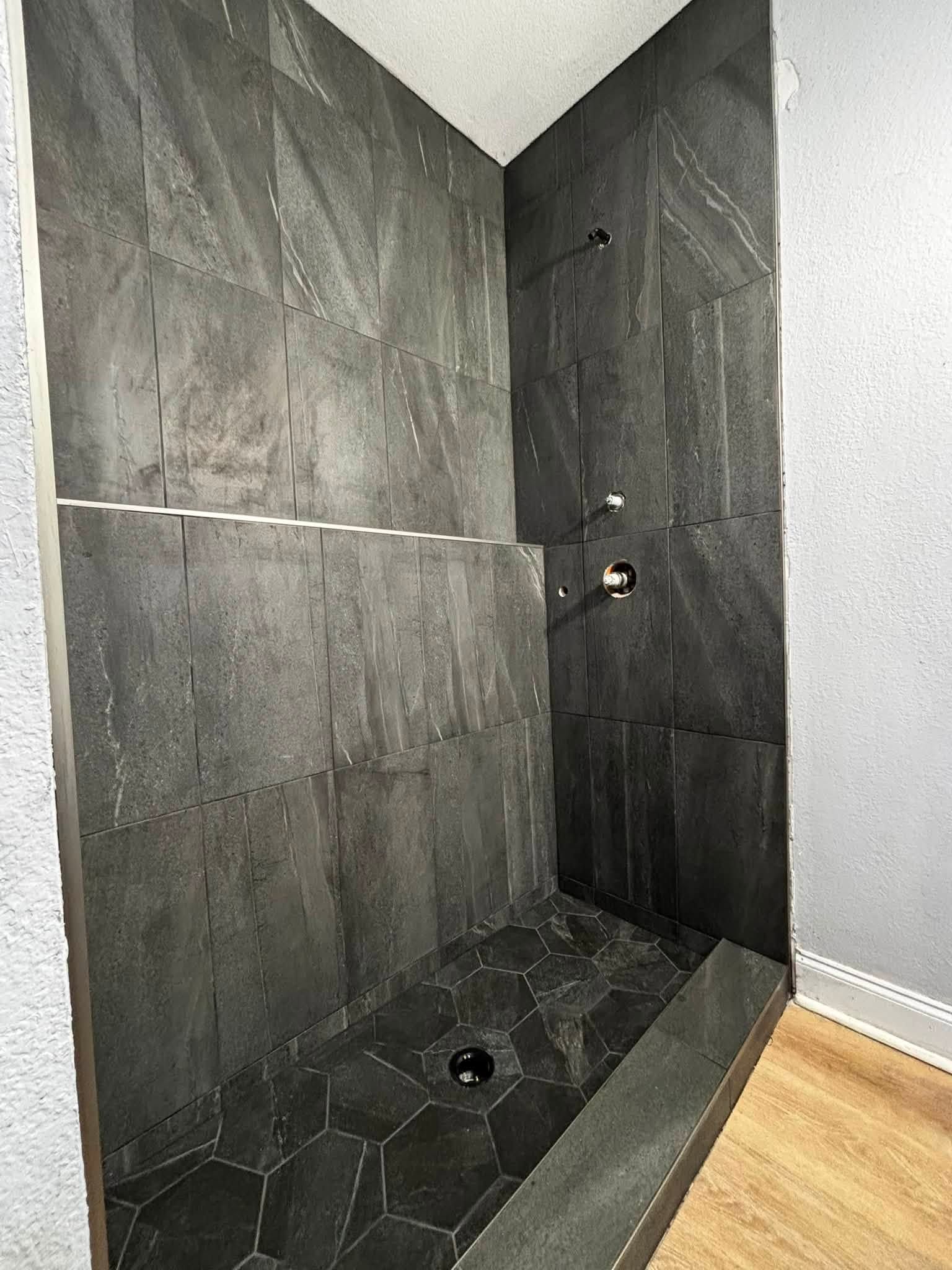 Black-tiled walk-in shower with hex tile floor, wall niche, and wood floor outside.