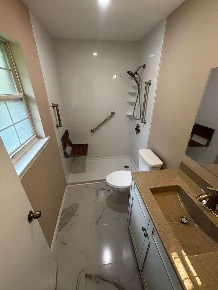 Small beige bathroom with toilet, shower, sink vanity, grab bars, and a window.