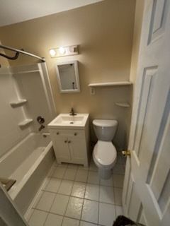 Small beige bathroom with shower-tub, sink, mirror, toilet, and tiled floor.