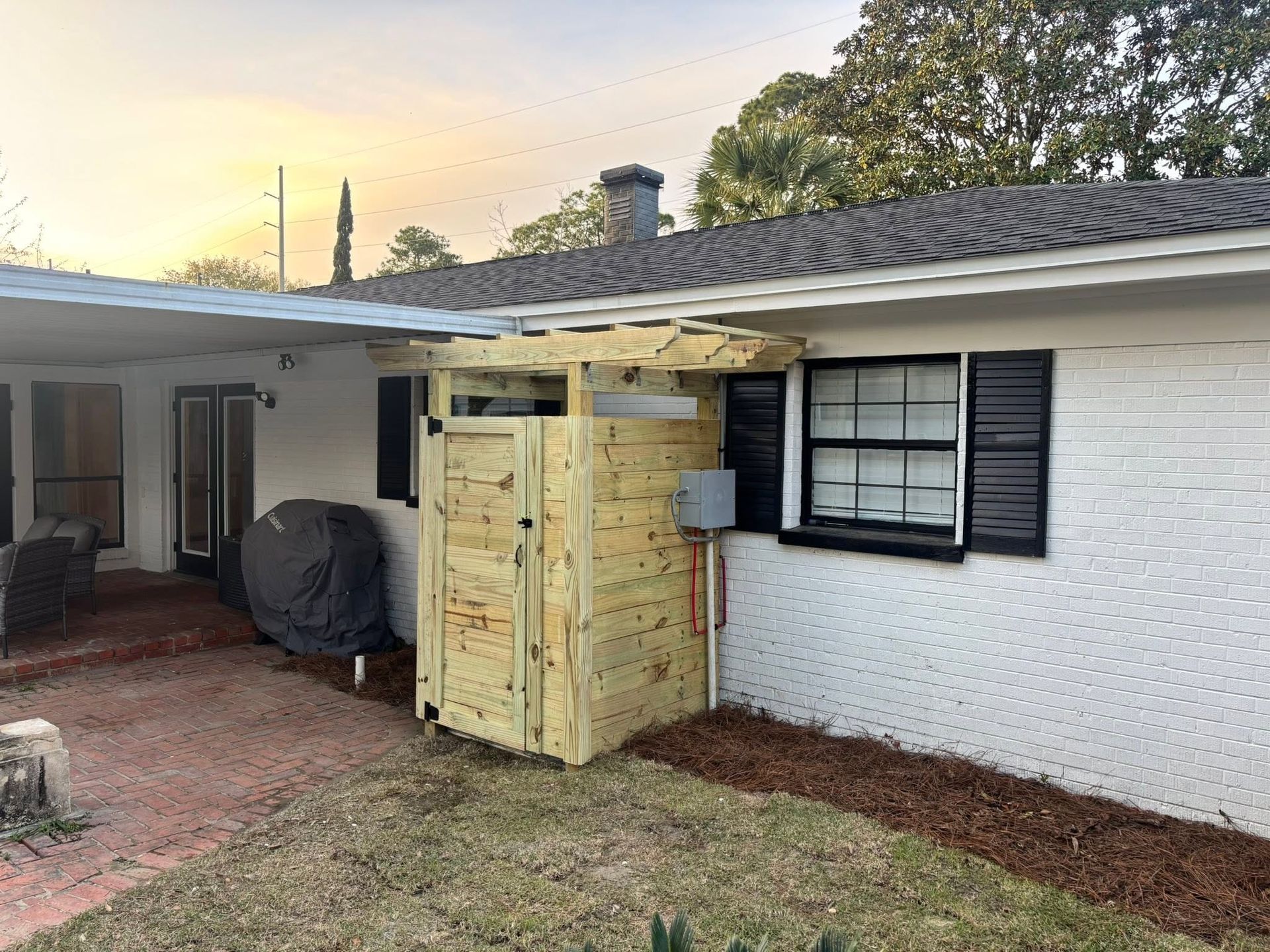 Partially built wooden shed beside a white house with a screened door and covered patio at sunset