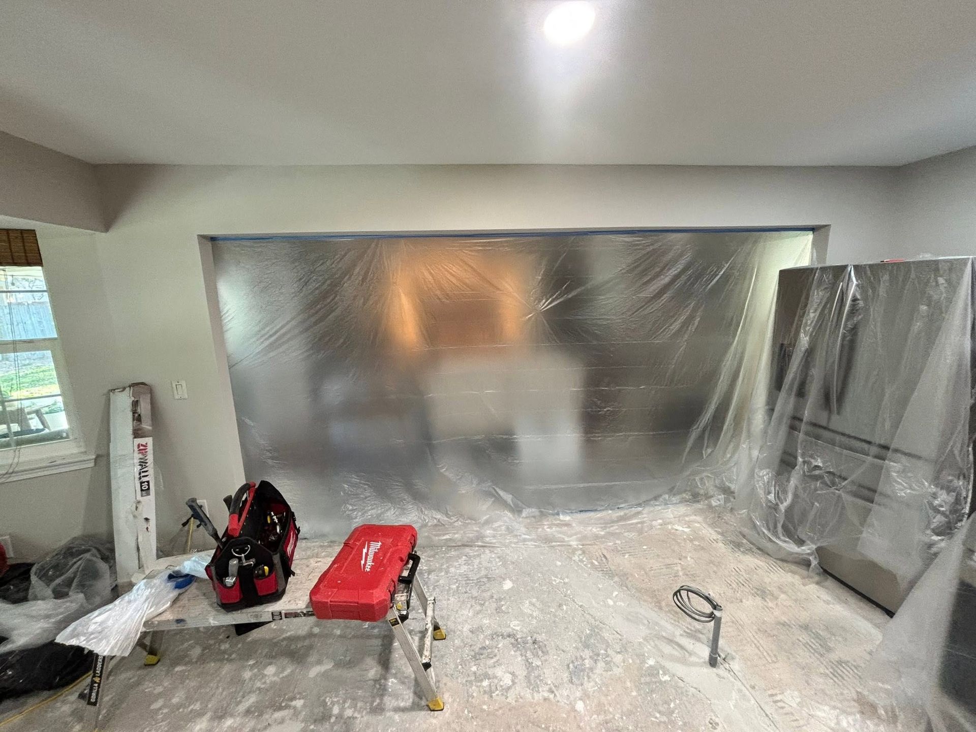 Room under renovation with plastic sheeting over walls and furniture, tools and supplies on the floor