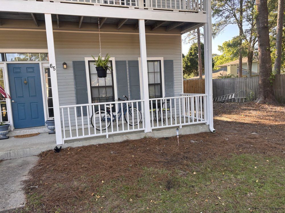 Gray house porch with white railing, blue door, and hanging plant in a yard.
