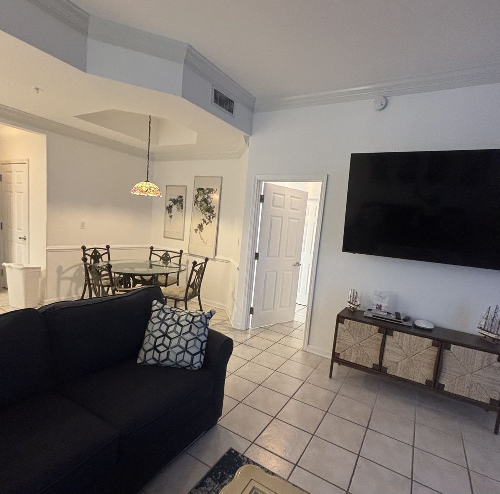 Open-concept living and dining room with black sofa, wall-mounted TV, tiled floor, and pendant lights.