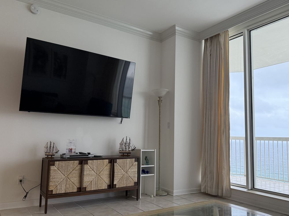 Bright living room with wall-mounted TV, console table, and large sliding glass door with curtains overlooking water.