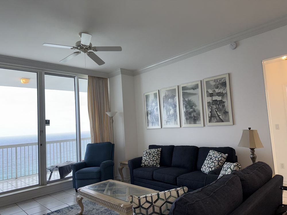 Bright living room with dark sofa, glass coffee table, ceiling fan, balcony view, and wall art.