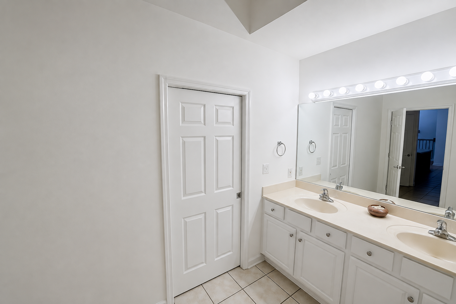 Bathroom vanity with large mirror, double sink, and a closed white door on tiled floor.