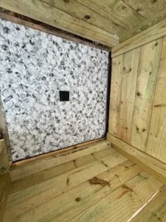 Wooden interior corner with a speckled stone wall and a small black square vent or outlet.