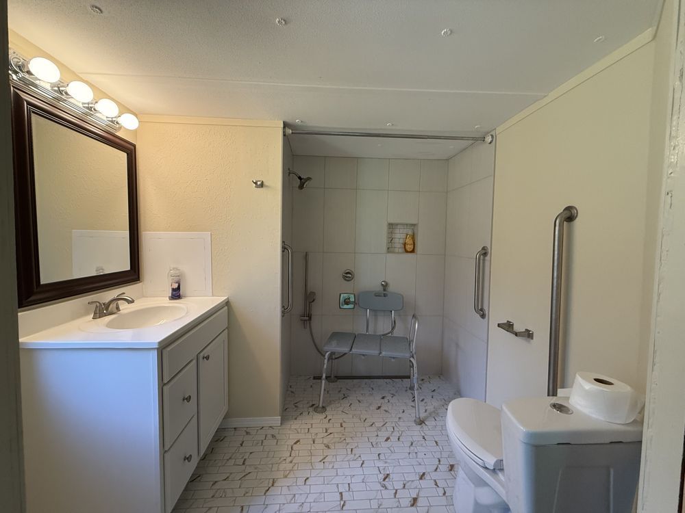 Bright bathroom with vanity, large mirror, walk-in shower, and toilet with grab bars.