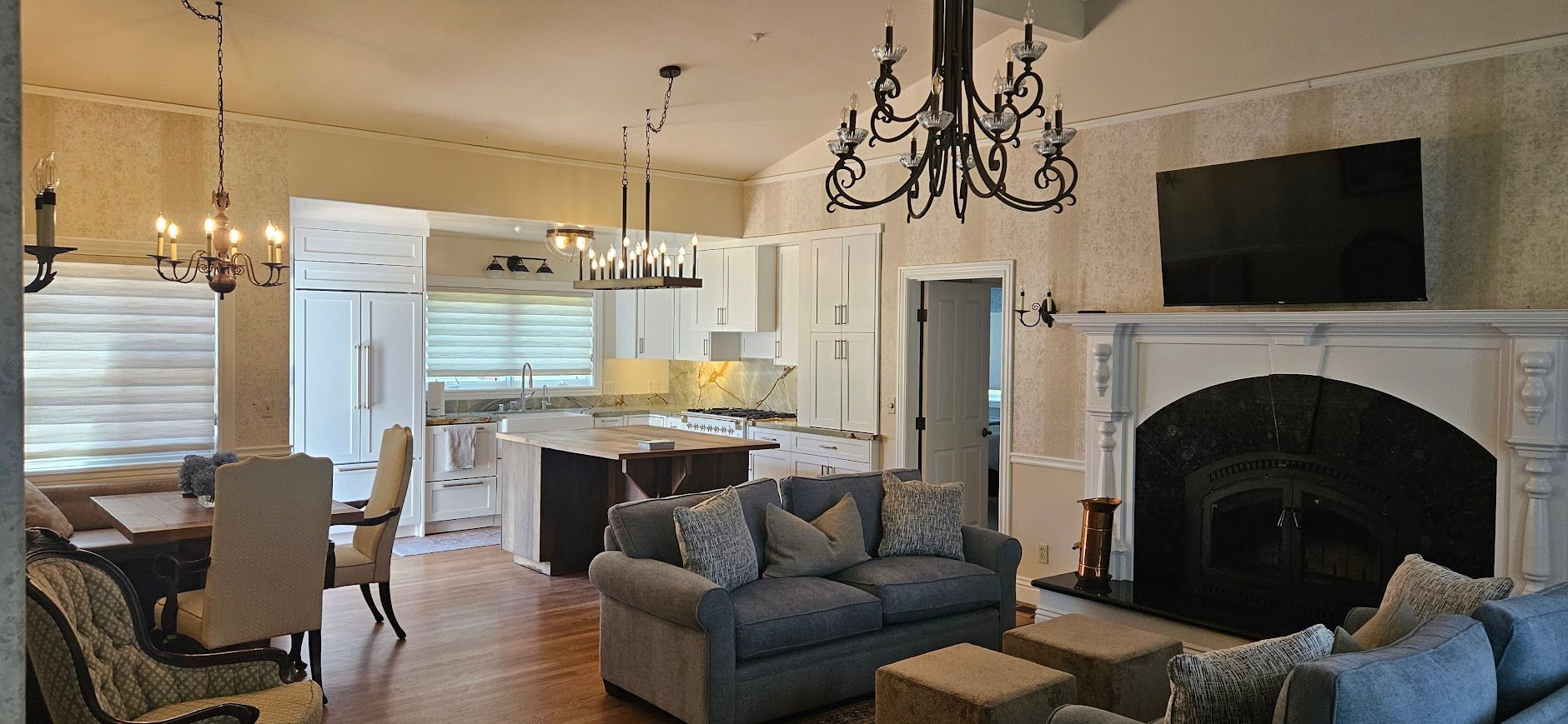 Spacious open-plan living room with gray sofa, large fireplace, chandelier, and kitchen in the background