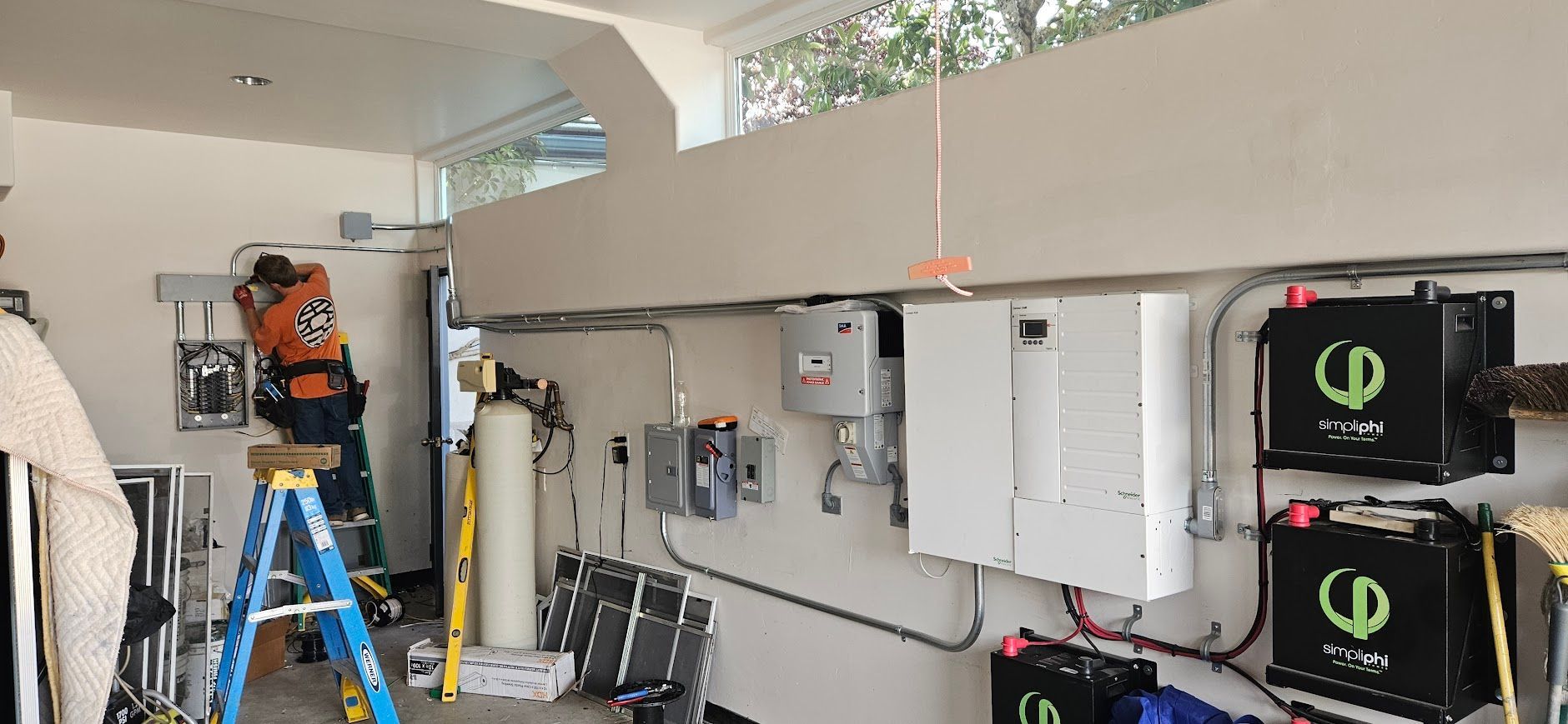Technician in orange safety gear works on electrical equipment in a bright utility room with wall-mounted boxes.