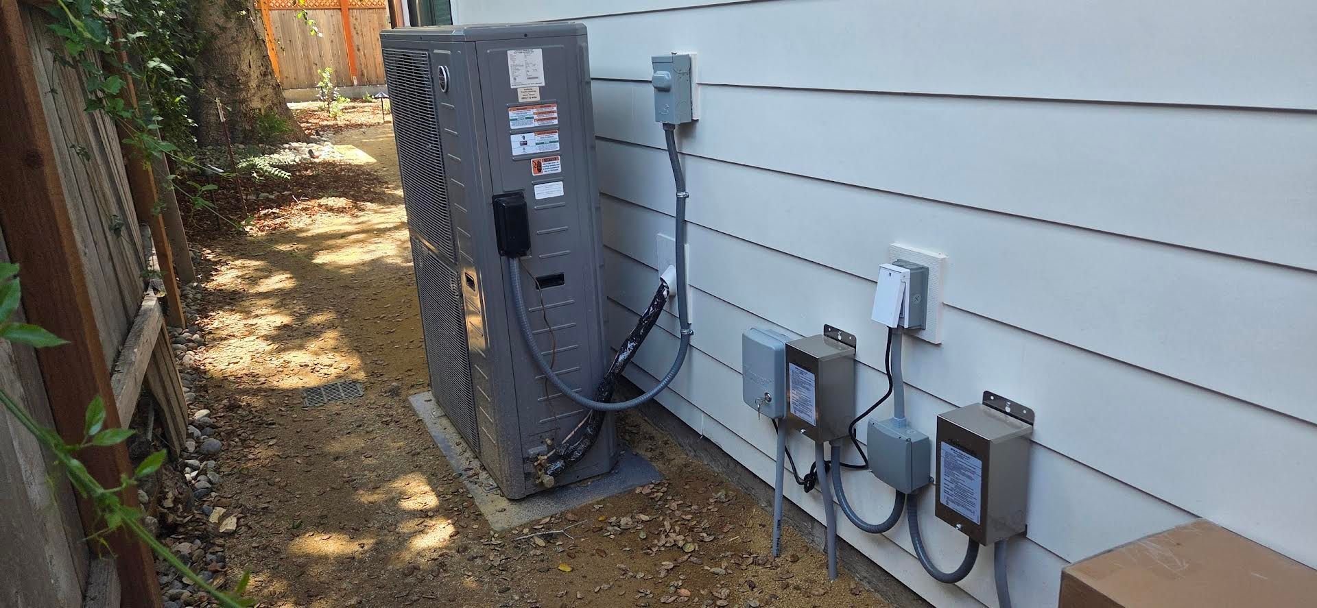 Outdoor HVAC unit and electrical boxes beside a house with gray siding and a narrow dirt path