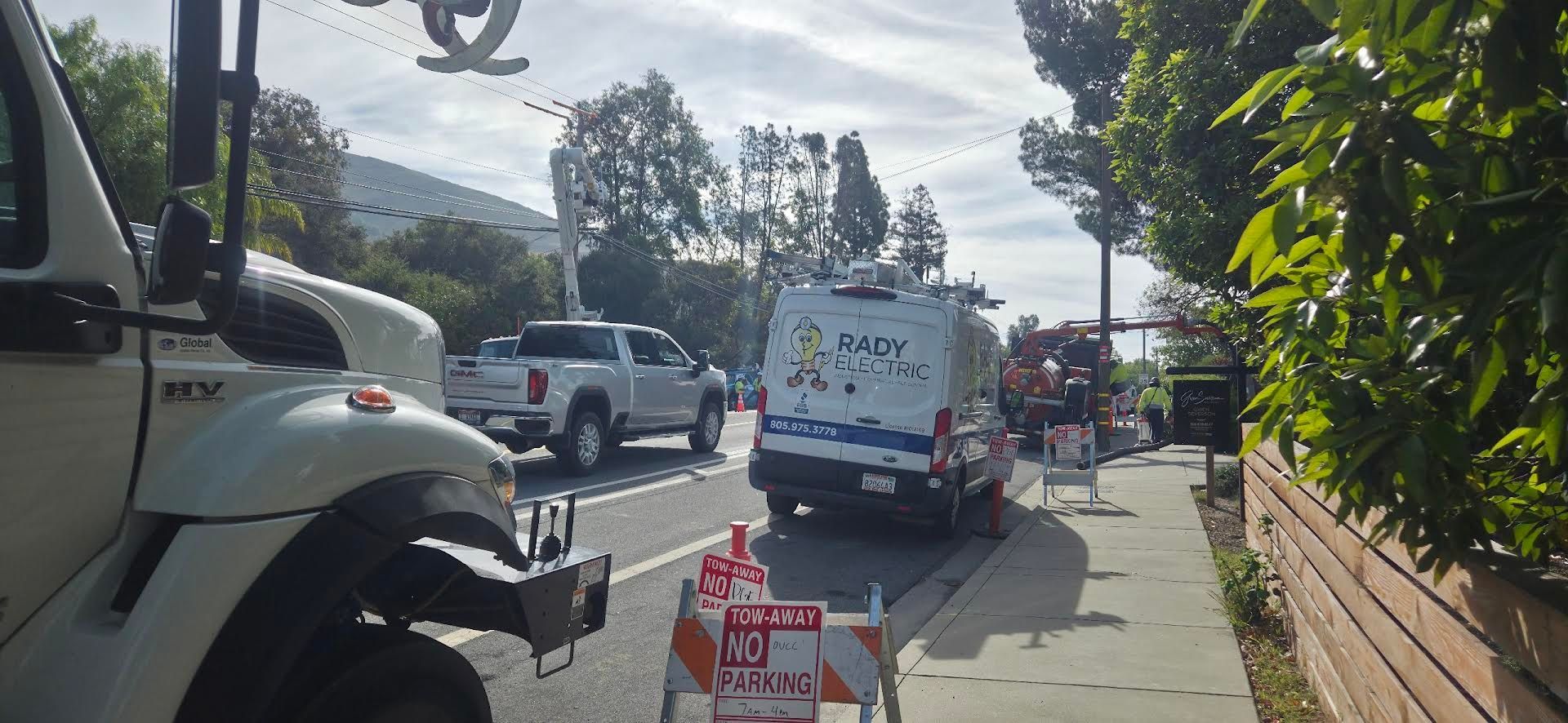Roadside sidewalk with parked trucks and a van under bright sun, next to a brick wall and trees