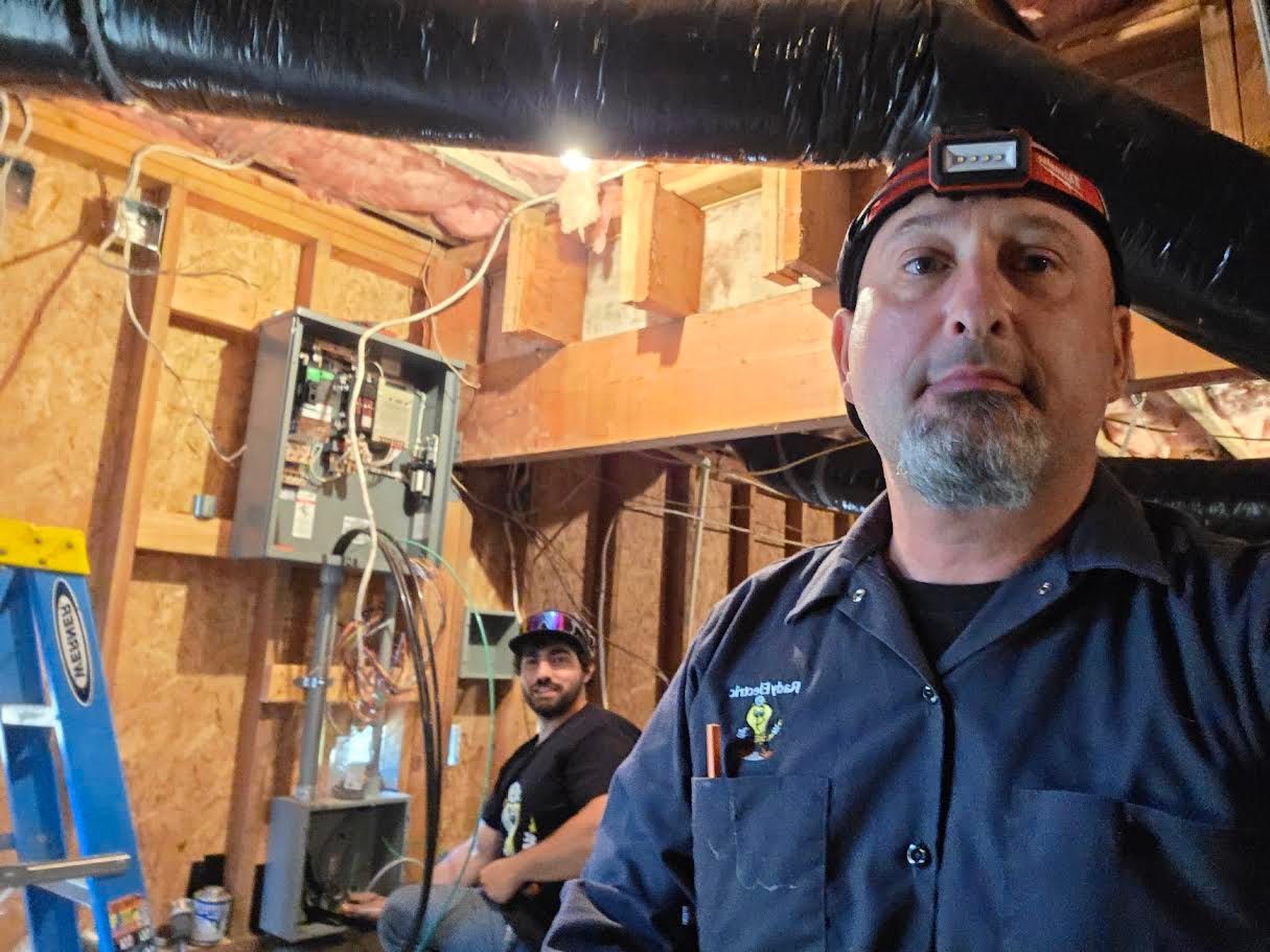 Two workers in a wood-framed attic with exposed pipes, one in the foreground wearing a headlamp.