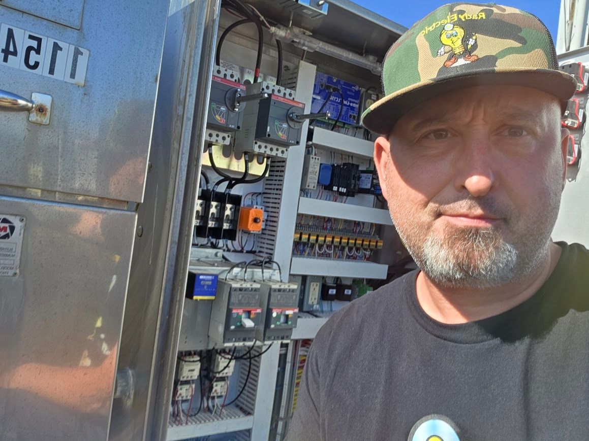 Man standing beside an open electrical panel in daylight, wearing a camouflage cap and black shirt.