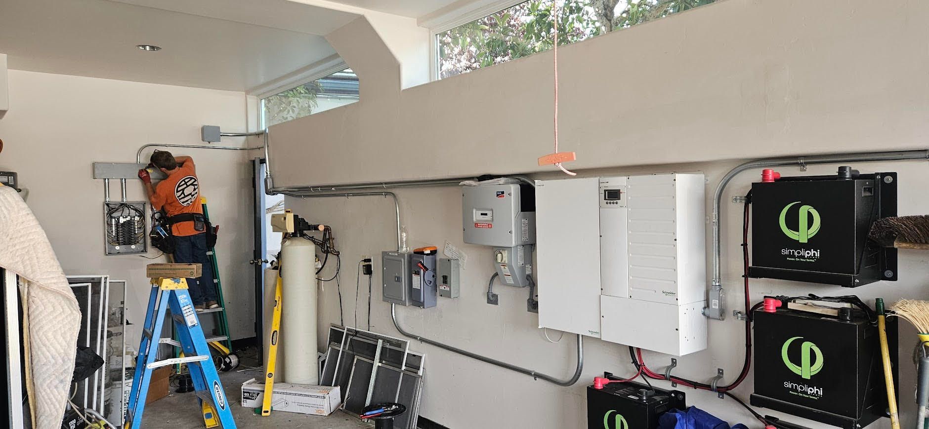 Technician on a ladder inspecting a wall-mounted electrical system in a garage with solar equipment.