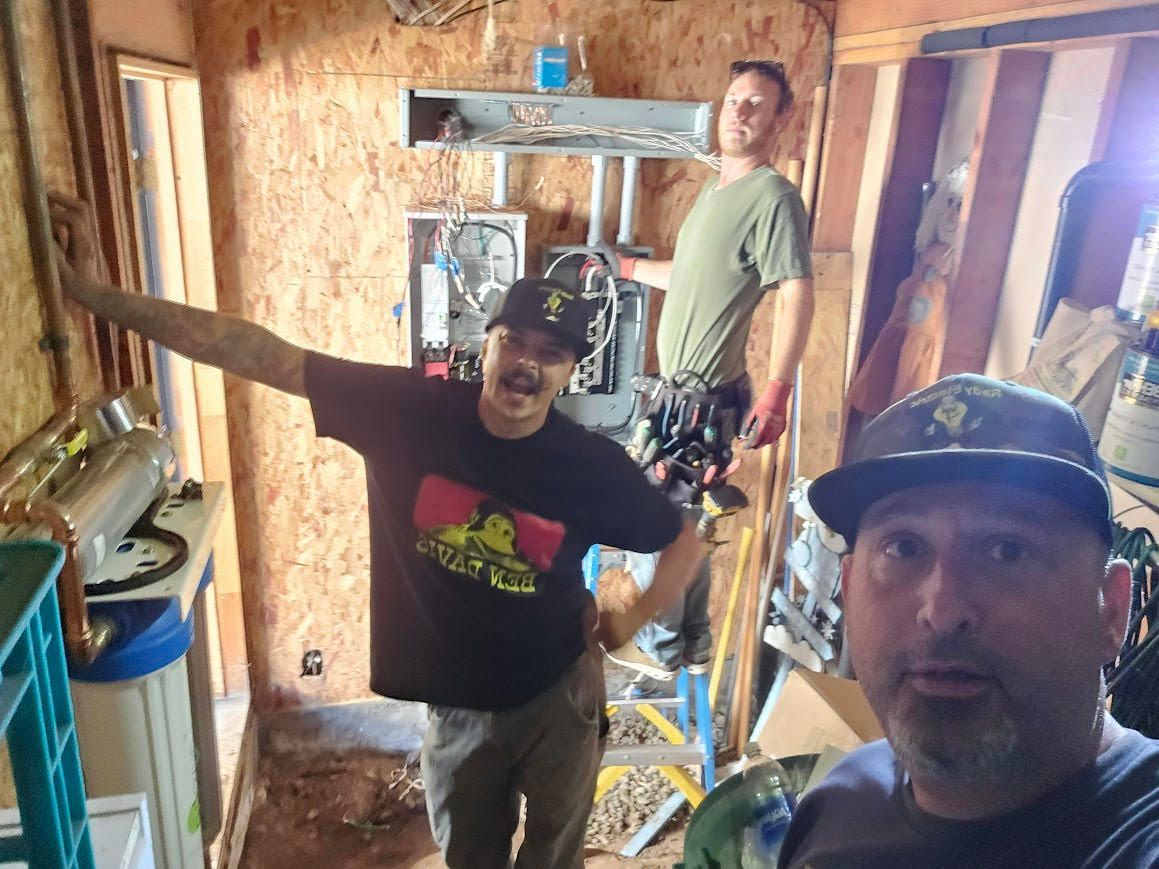 Three people smiling in a small workshop, one taking a selfie in the foreground.