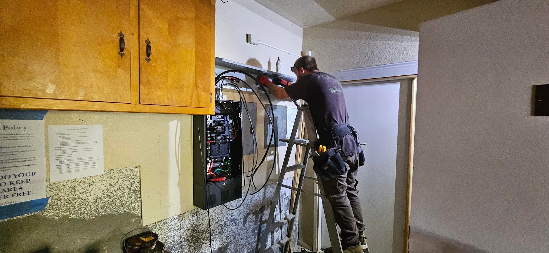 Worker repairing electrical panel in a hallway, wearing safety gear and reaching into open wiring.