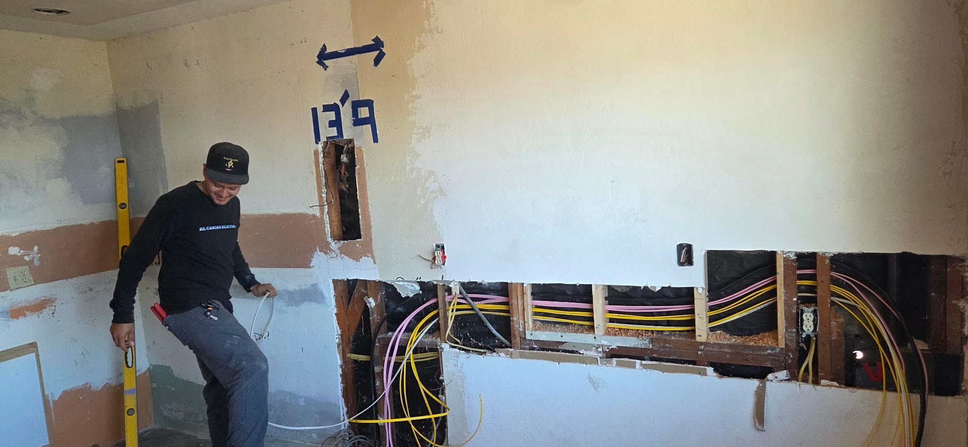 Worker inspecting exposed wiring in a damaged wall inside a room, with tools and debris visible.
