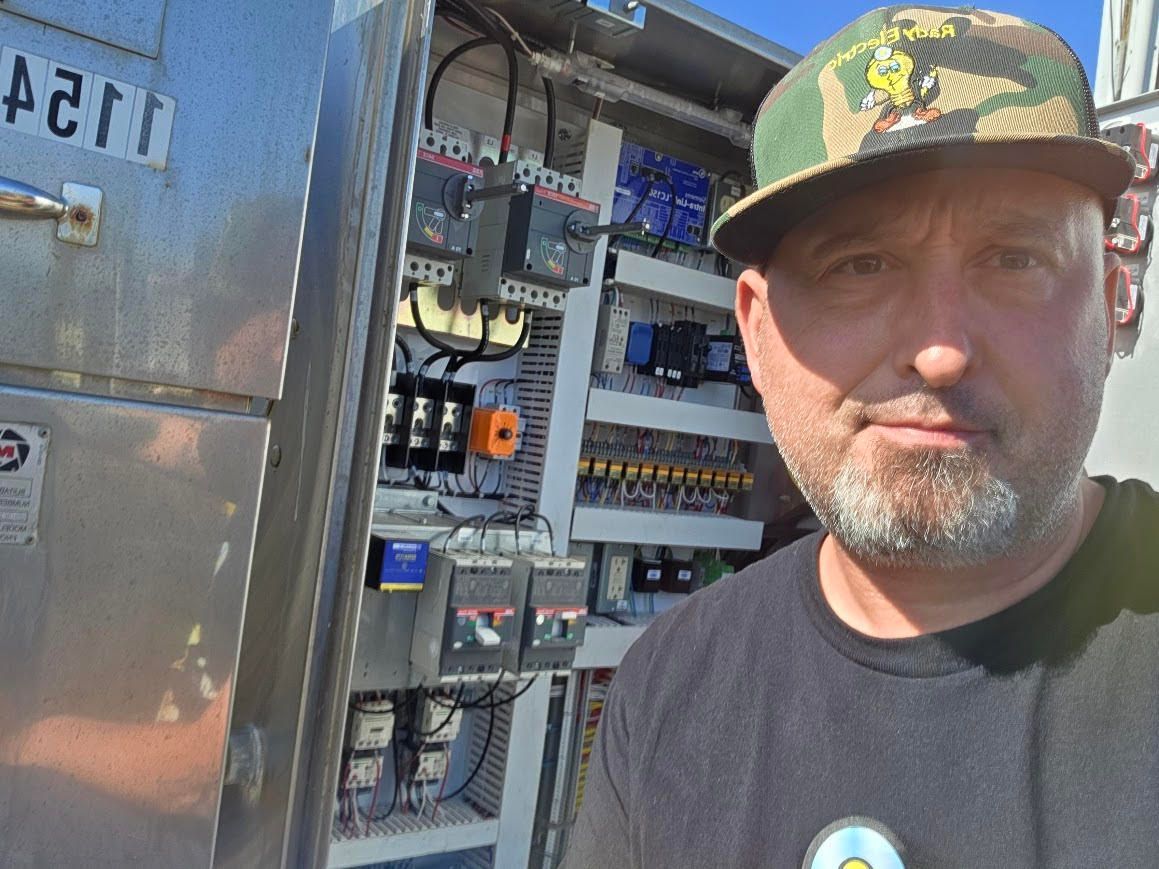 Man in a camouflage cap stands beside an open electrical panel outdoors.