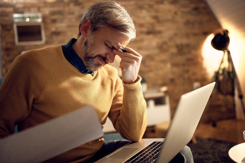 Man in a mustard sweater looks stressed while working at a laptop in a warm, brick-walled room.