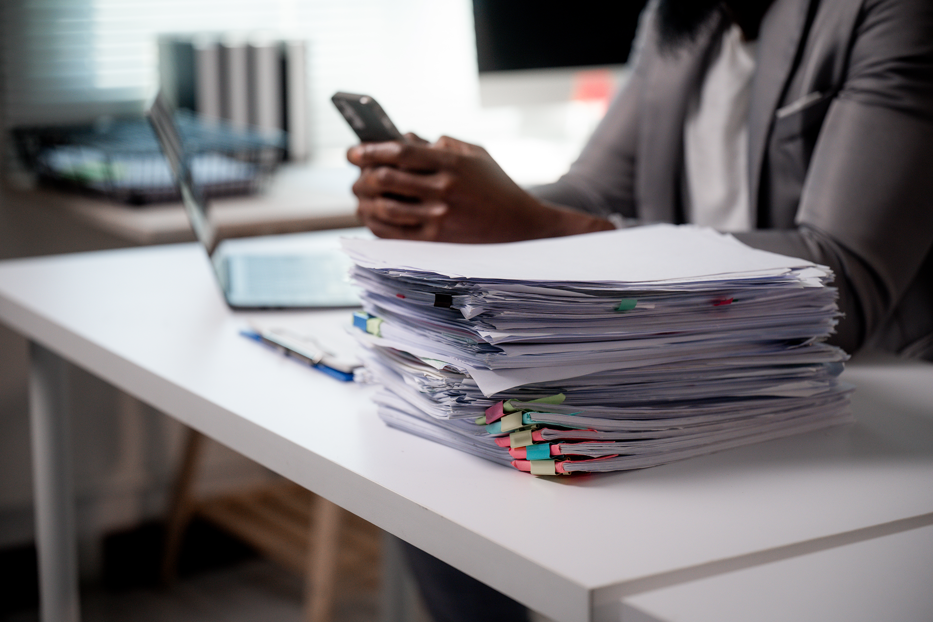 Person holding a phone beside a stack of paperwork on a white desk in an office