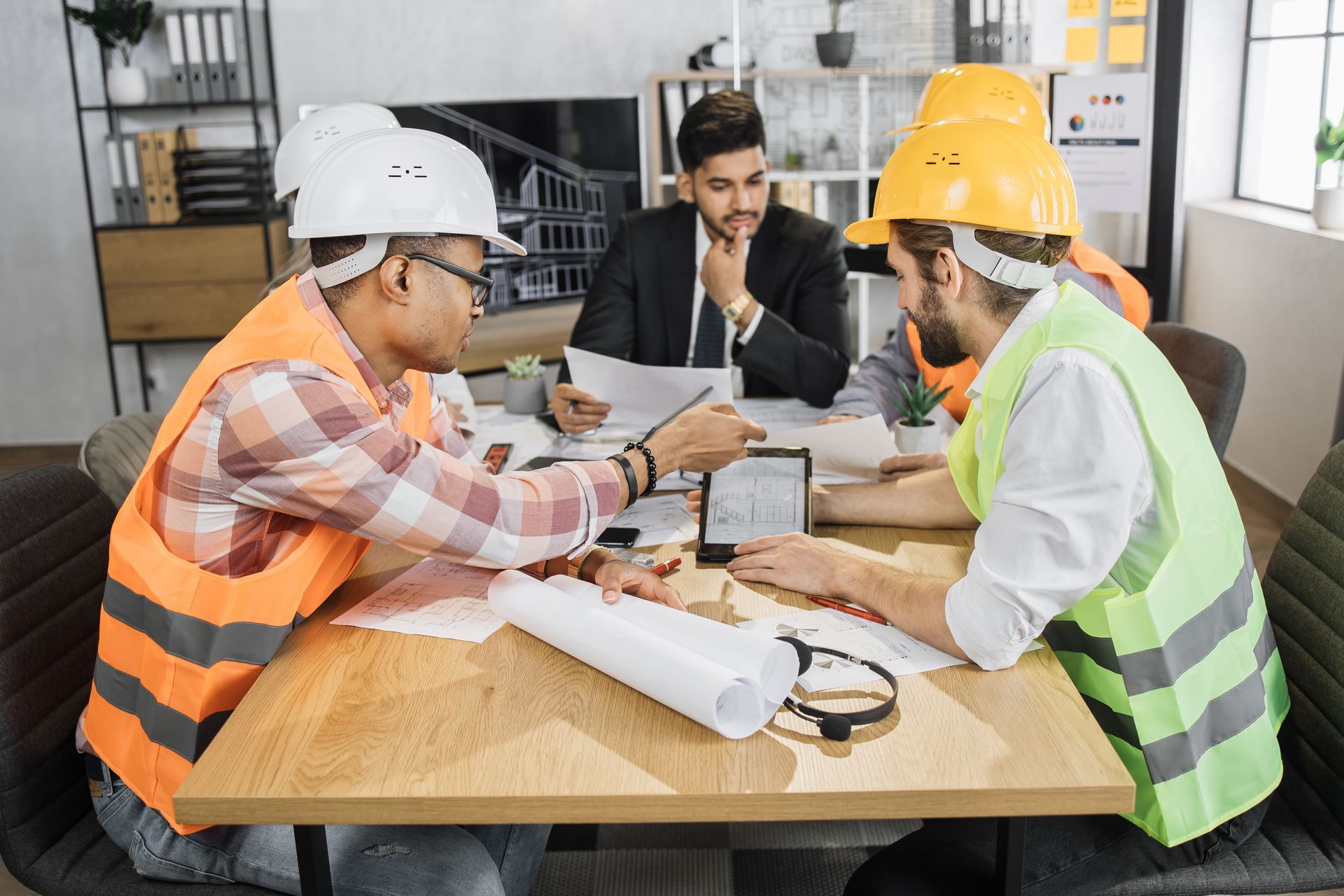 Three workers in safety vests and hard hats review blueprints at a table in an office.
