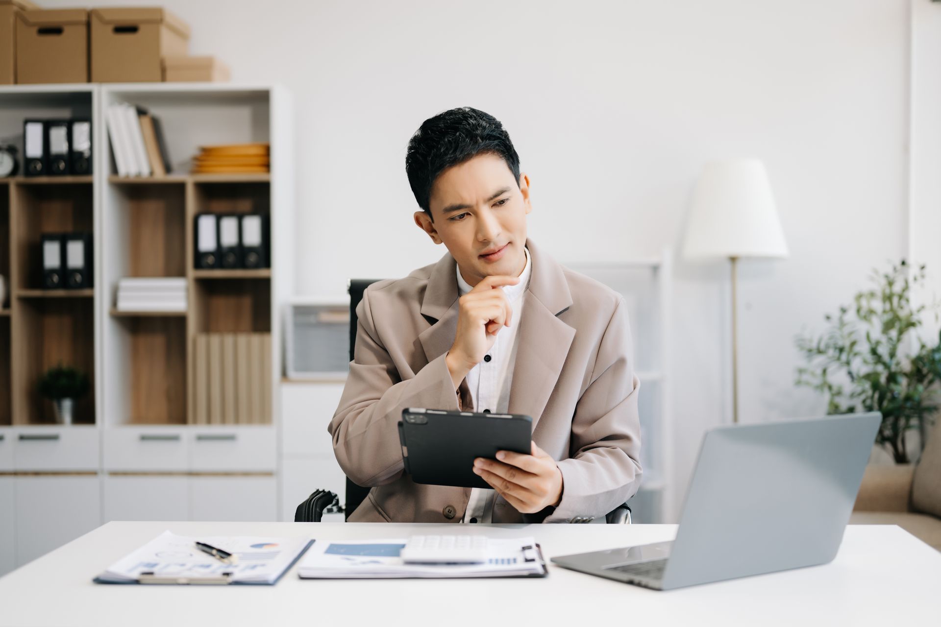 Person in a beige blazer reviewing a tablet at a desk with a laptop in a bright office
