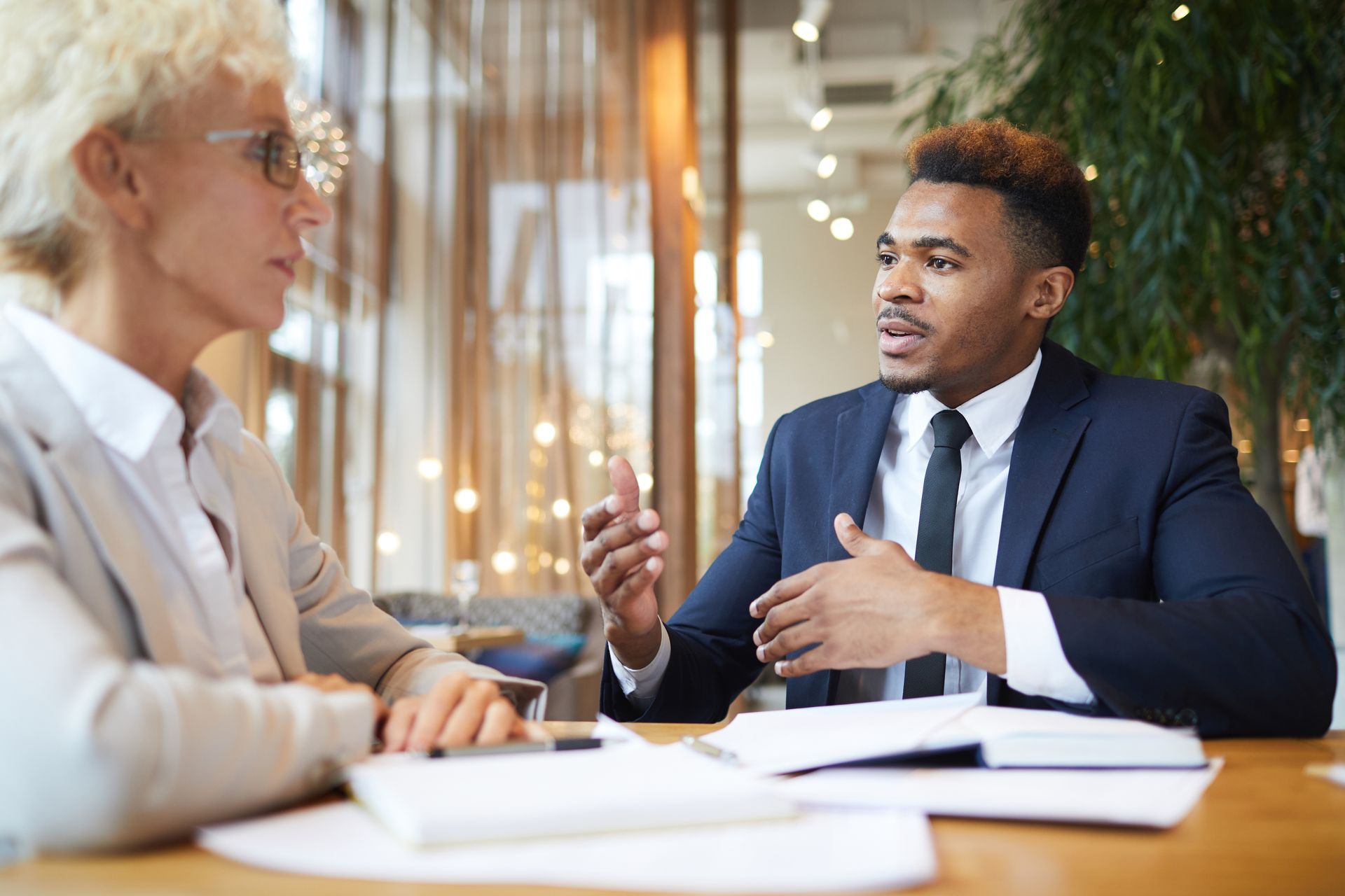 Two professionals in a meeting, speaking across a table with documents in a bright office.