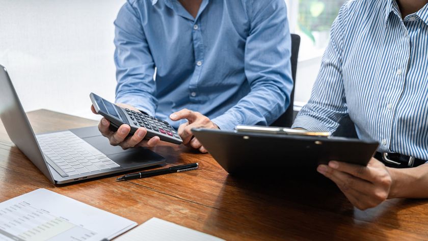 Two people at a desk using a calculator and tablet beside a laptop and papers