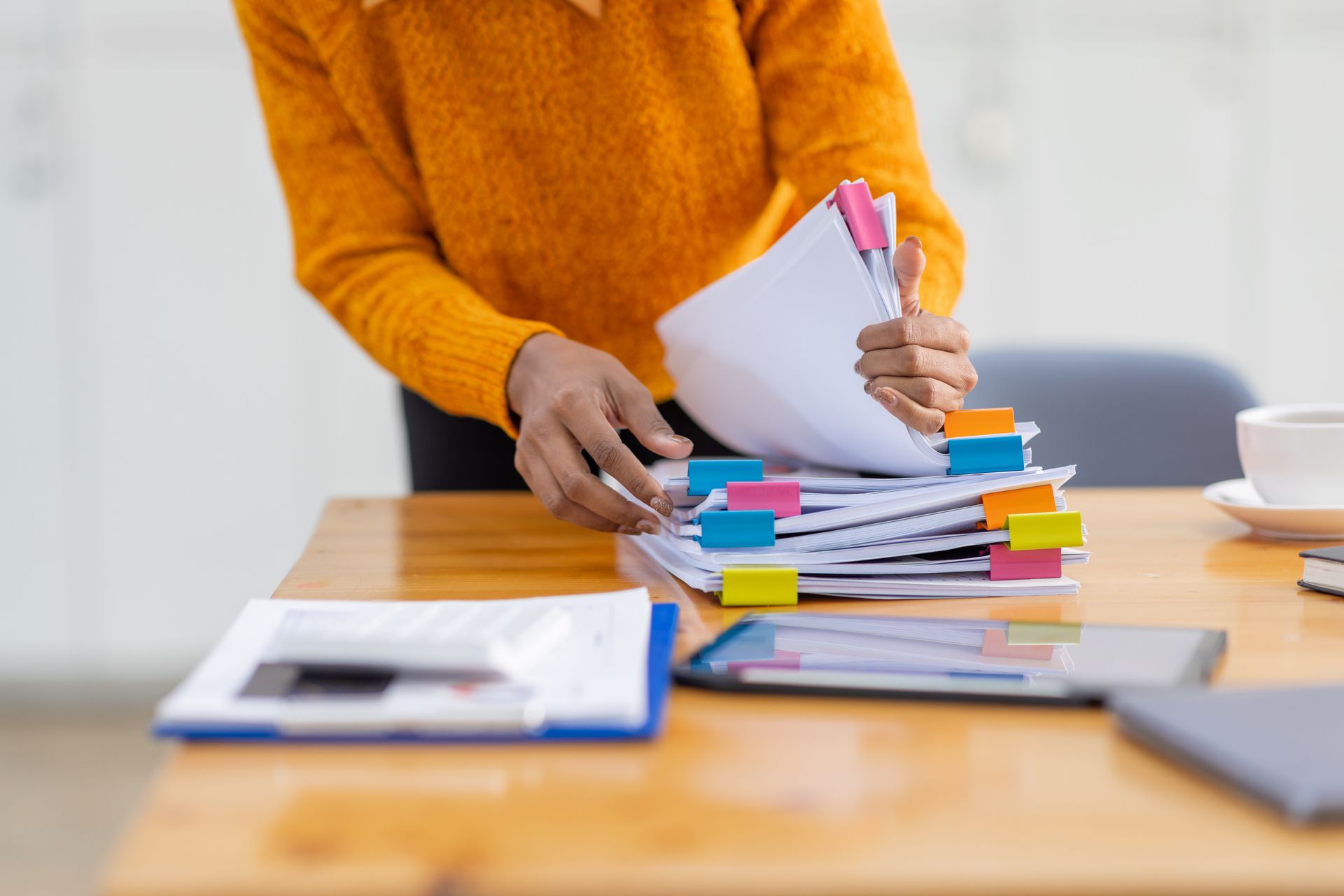 Person in orange sweater organizing a stack of papers with colorful sticky tabs on a desk