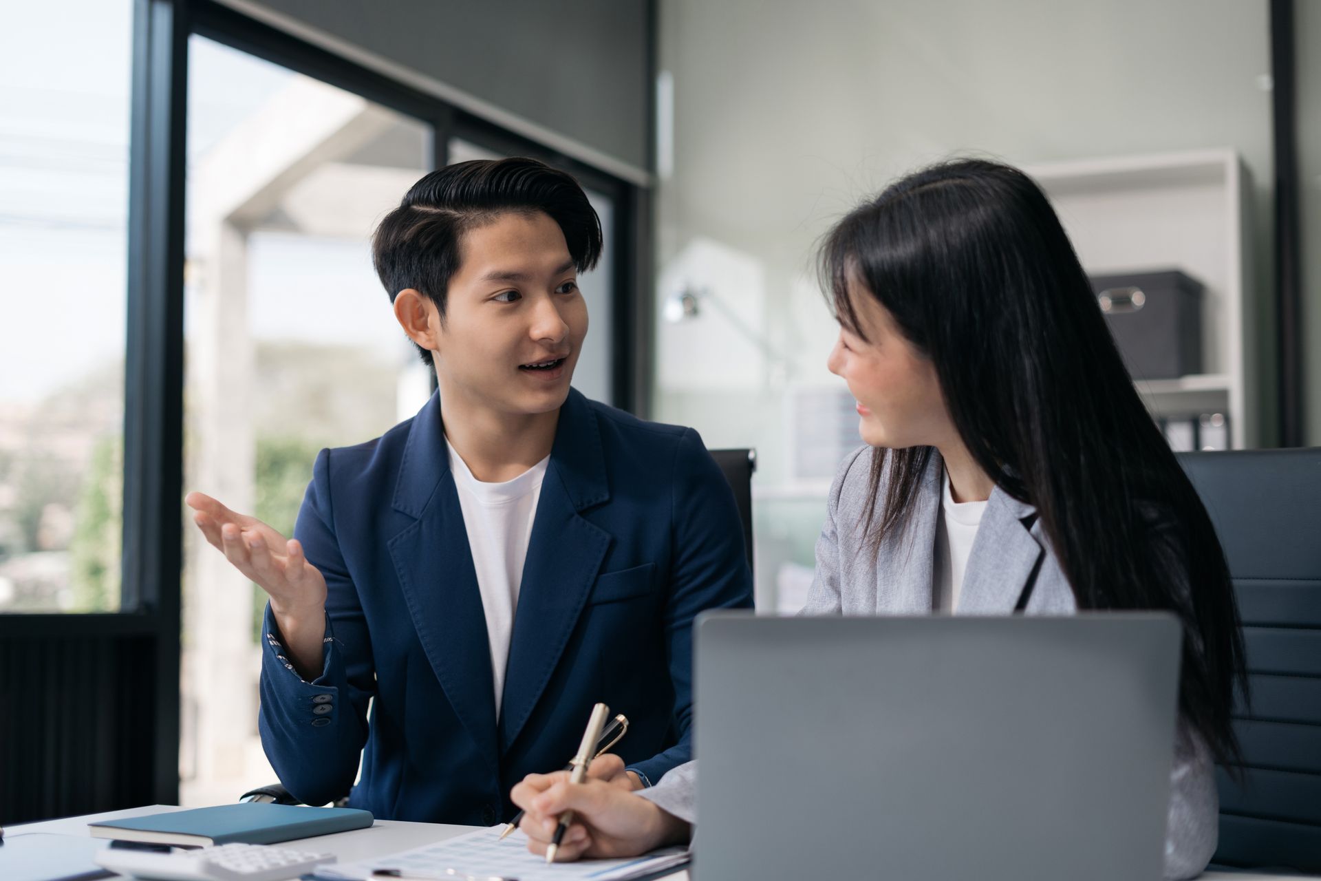 Two coworkers discussing at a desk beside a laptop in a bright office