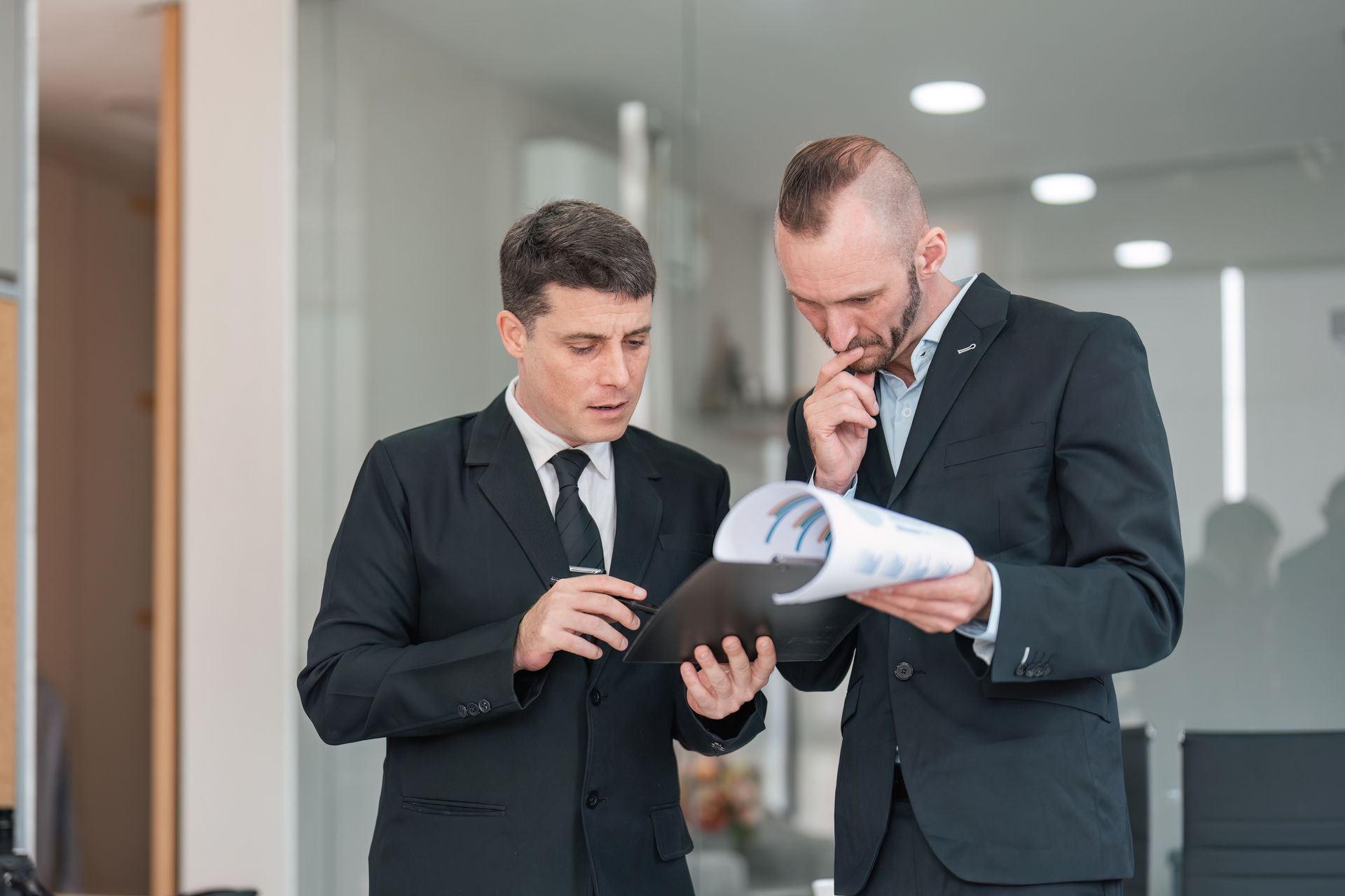 Two professionals in suits reviewing documents and a tablet in an office hallway