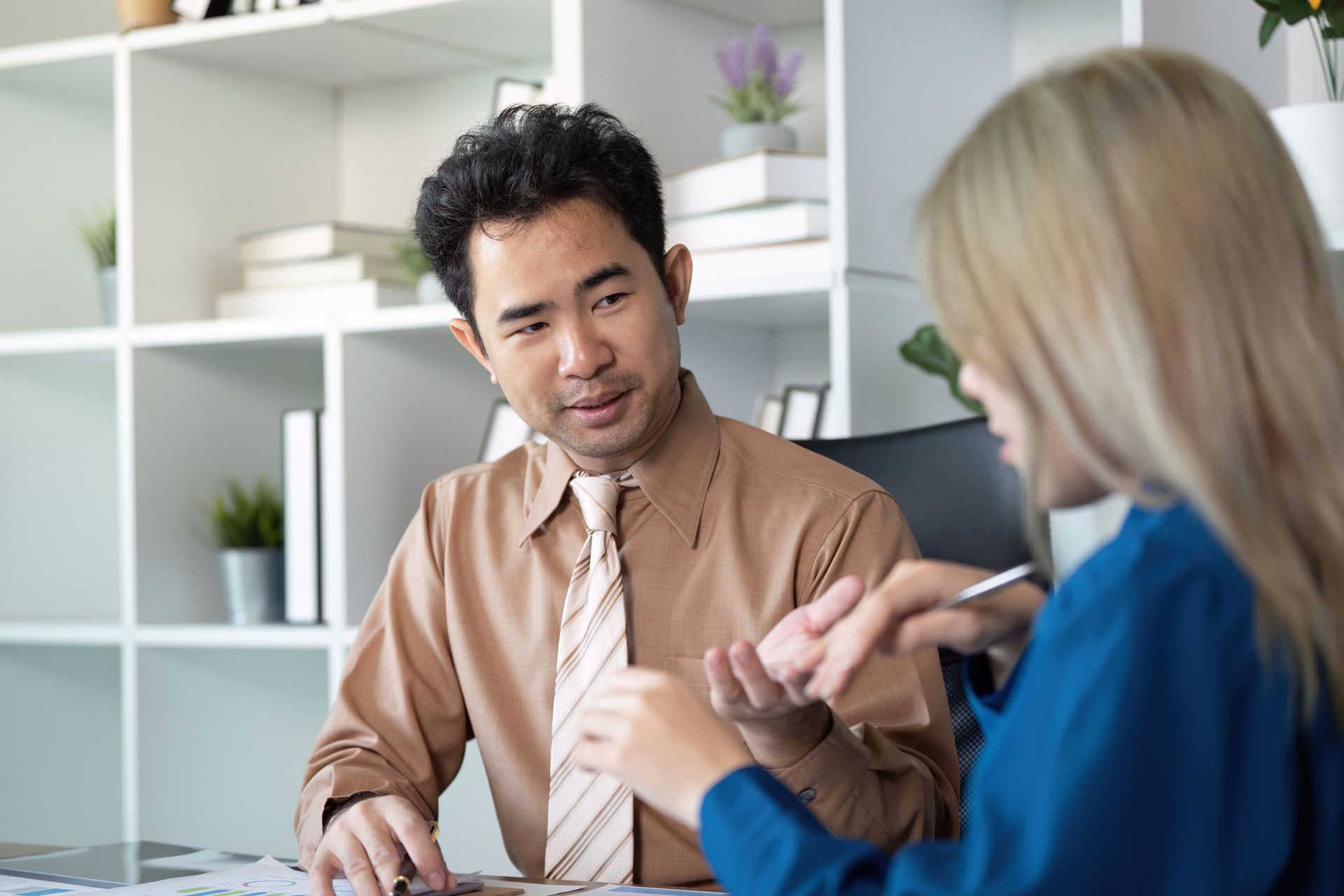 Two coworkers discussing paperwork at a desk in a bright office, one gesturing while the other listens