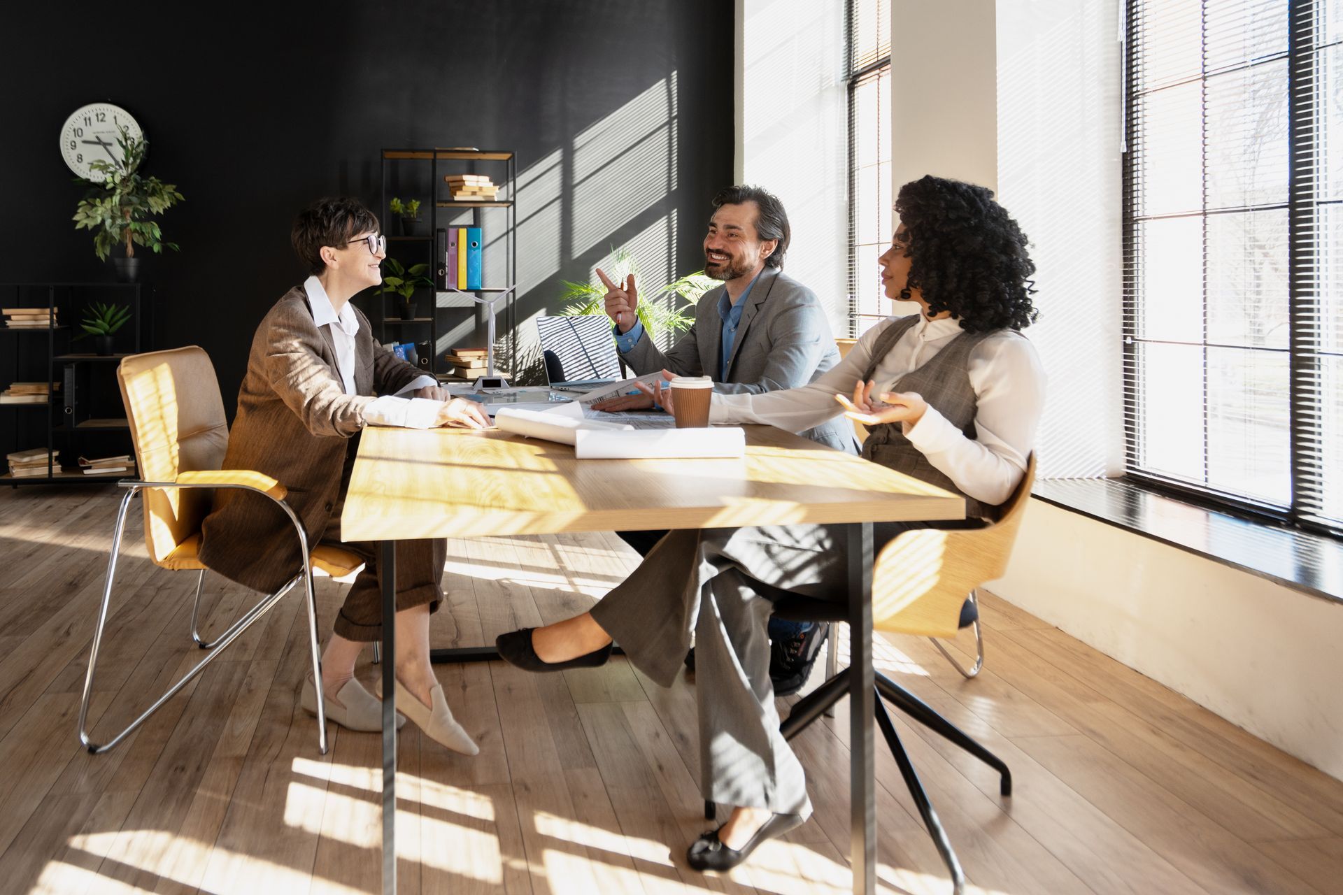 Three coworkers meeting around a table in a bright office, discussing documents and a laptop