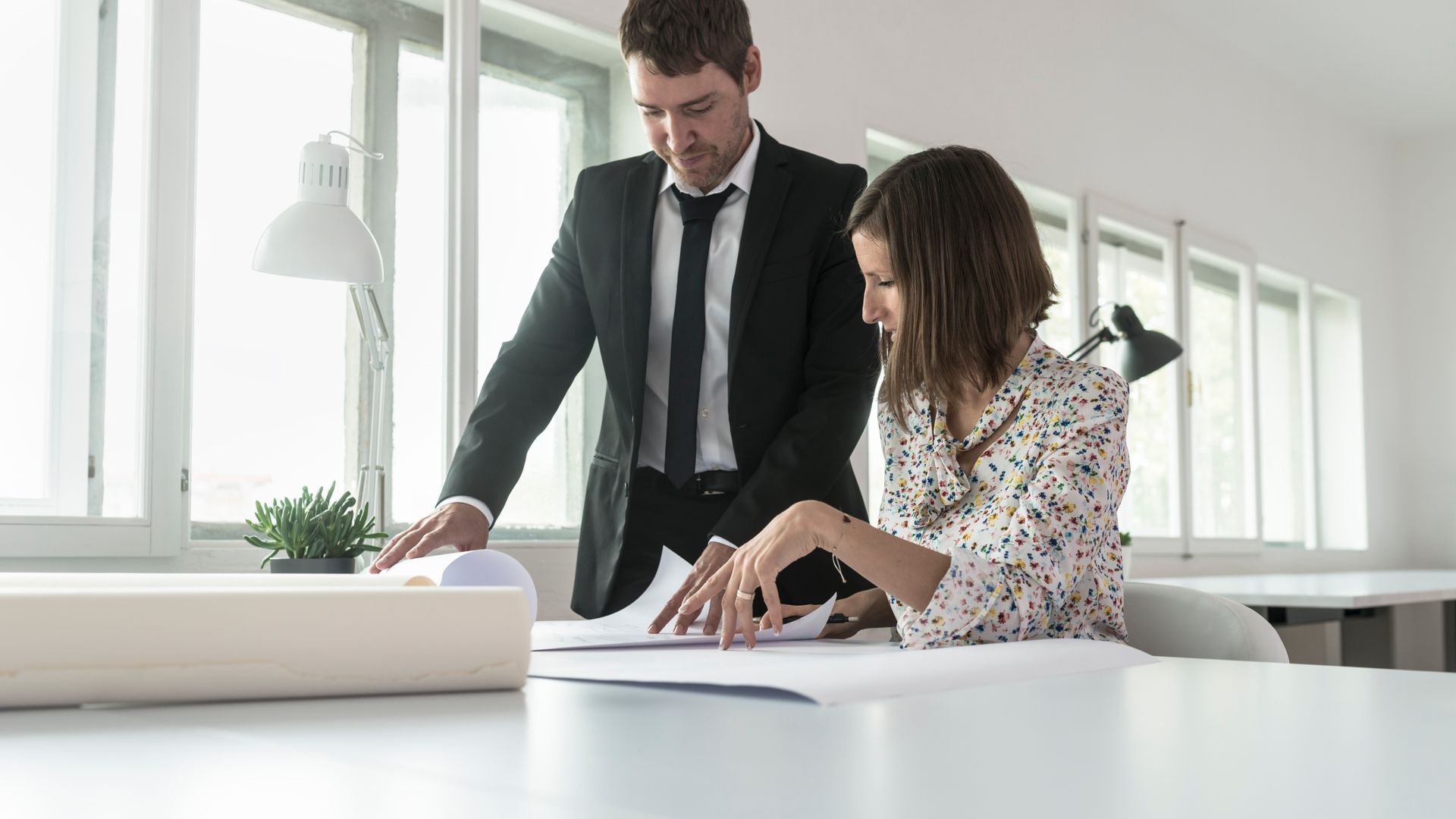 Two coworkers review documents at a bright office desk, one standing and pointing, the other seated and writing.