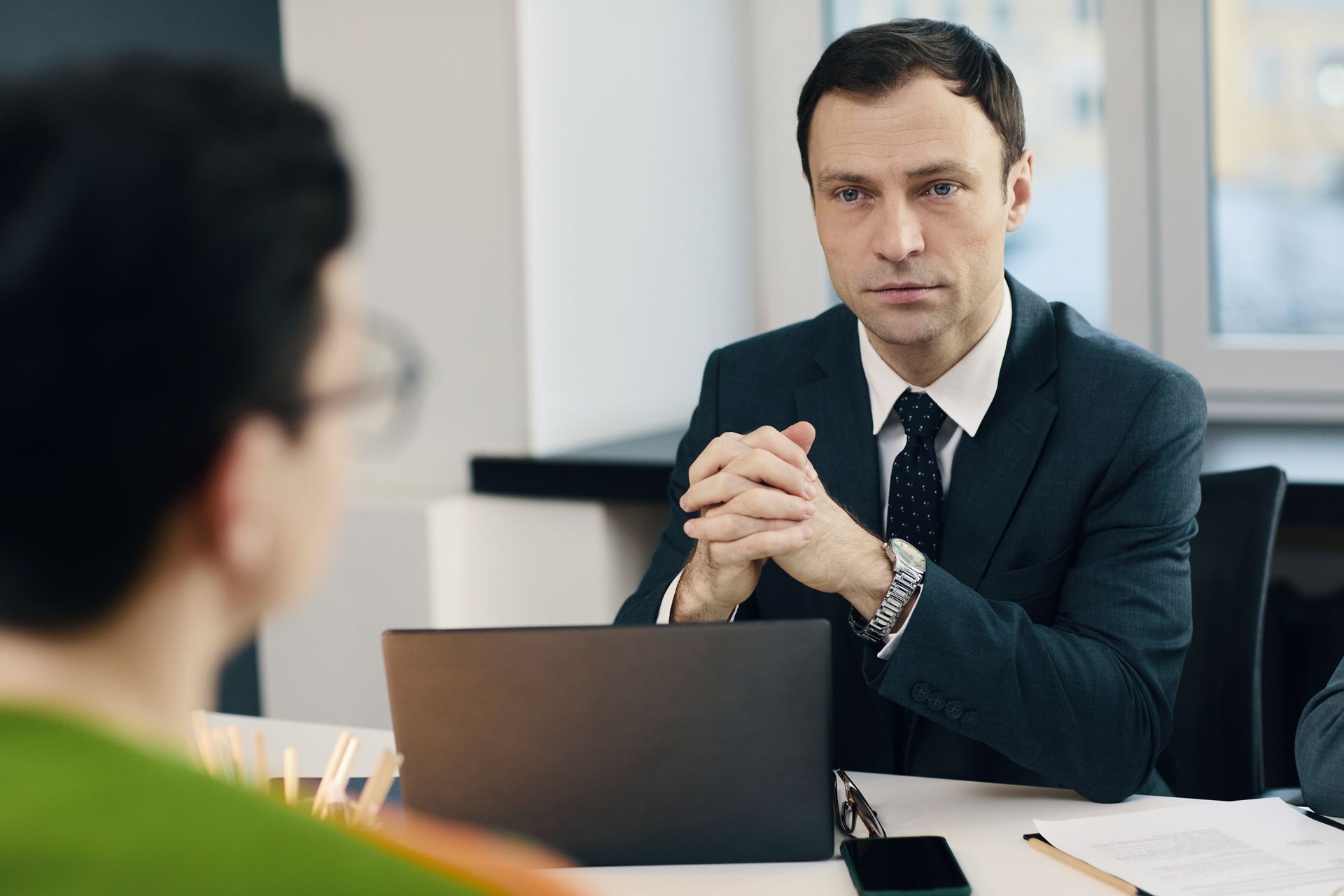 Two people in a business meeting, with a man in a suit speaking across a laptop in a bright office.