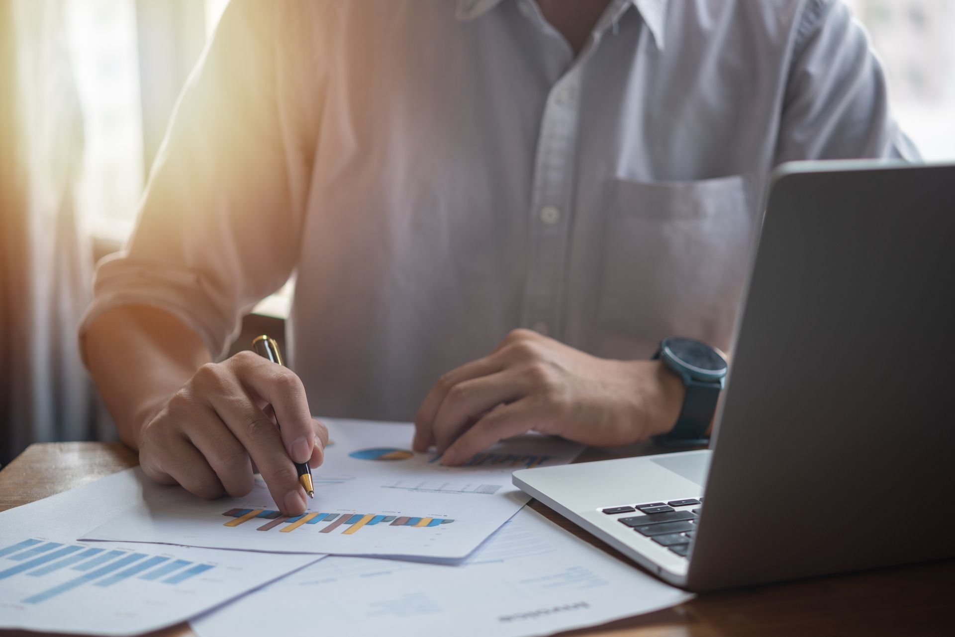 Person reviewing charts with a pencil beside a laptop in a sunlit office