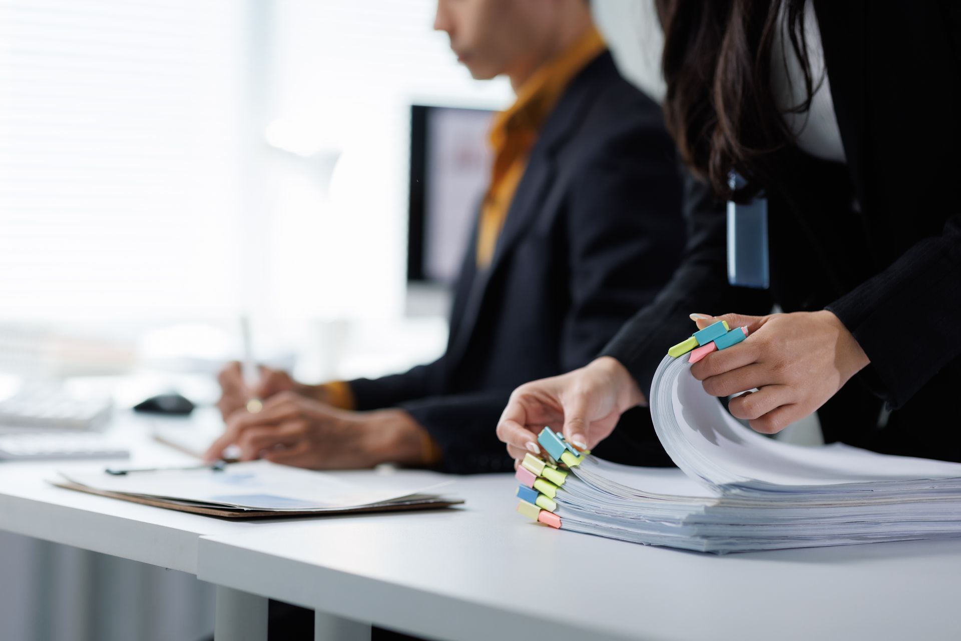 A person in a business suit sorts through a stack of documents organized with colorful index tabs at an office desk.