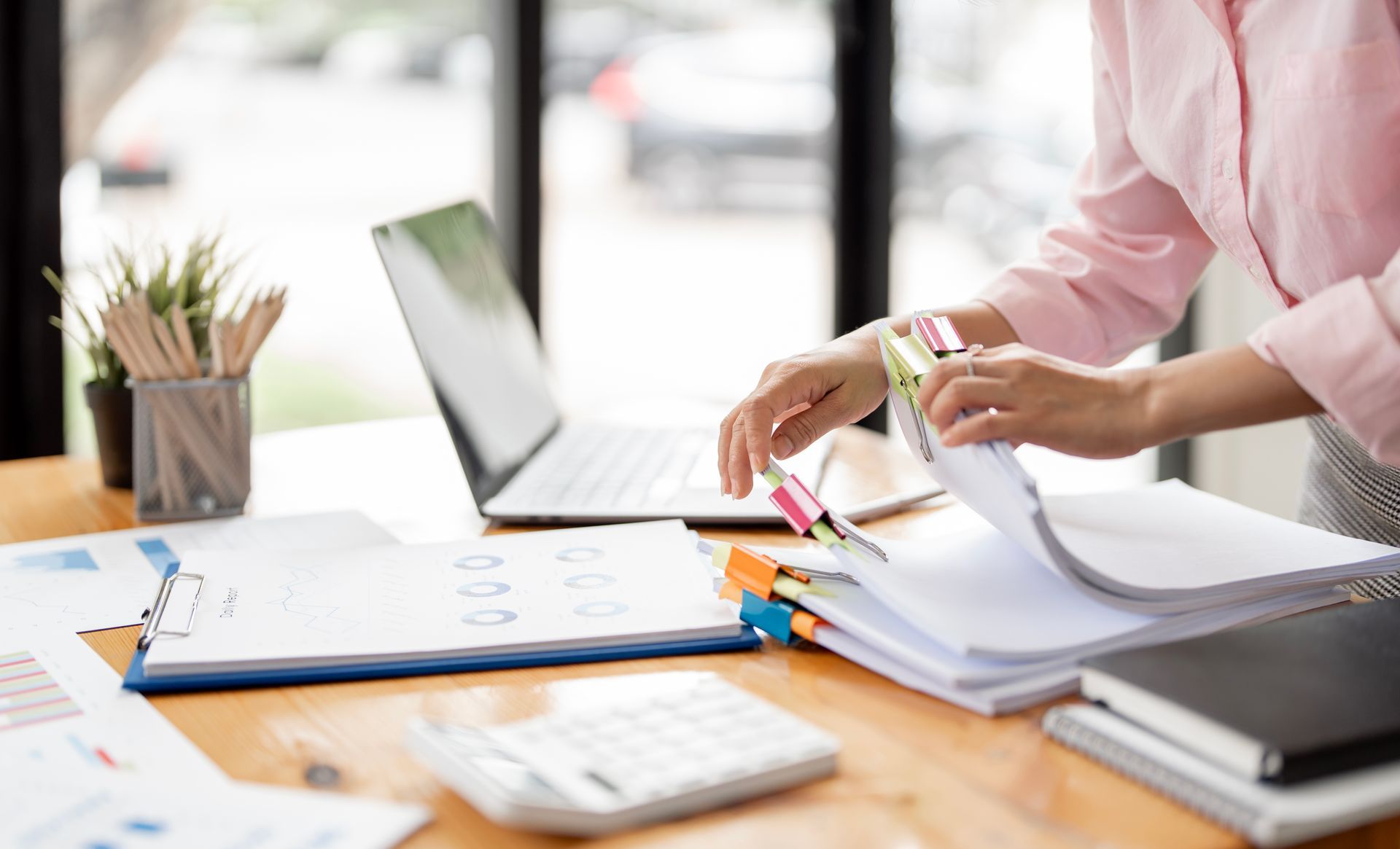 Person organizing papers at a desk with laptop, calculator, and office supplies in a bright workspace