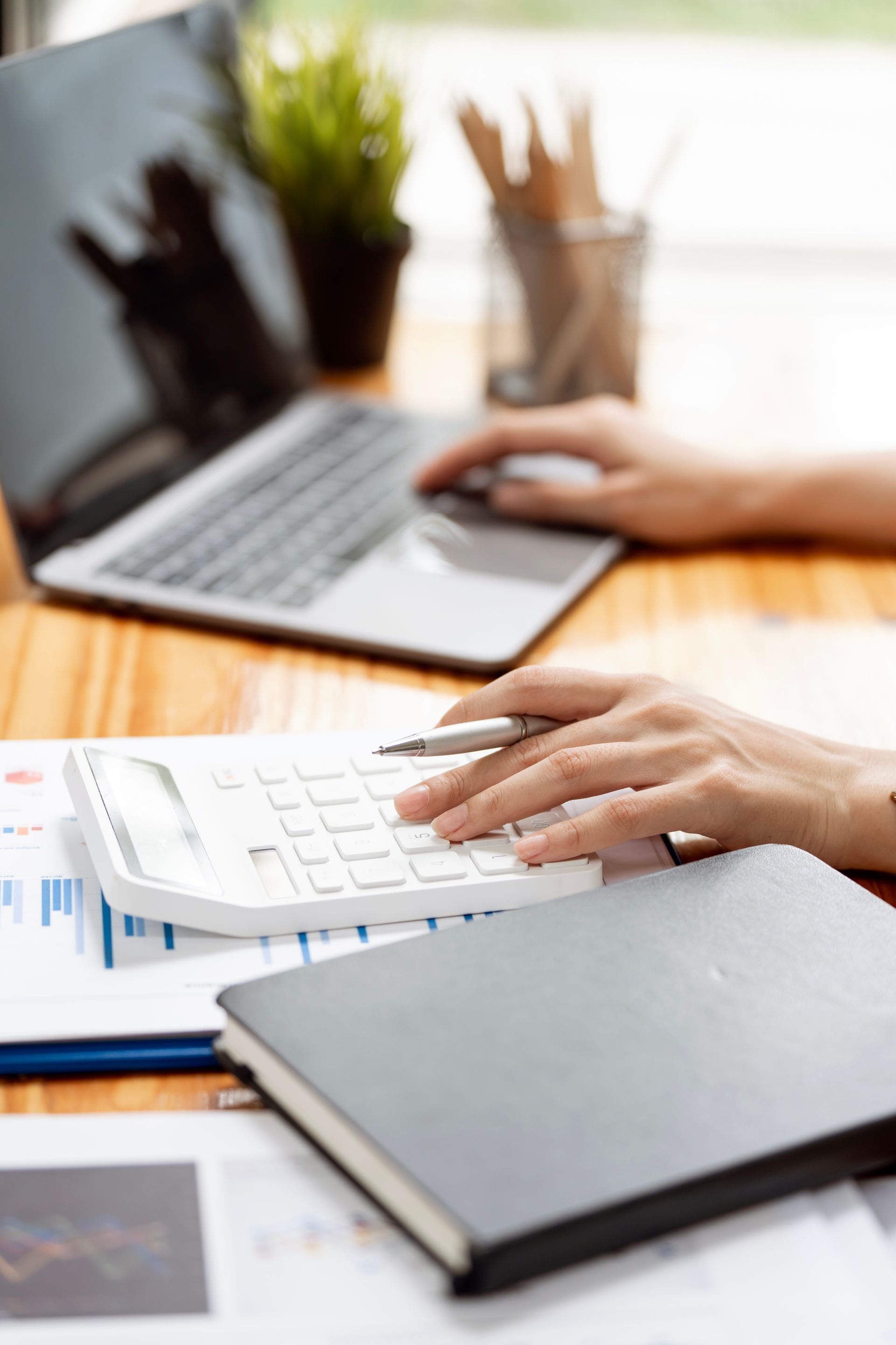 Hands typing on a white keyboard beside a notebook and laptop on a bright desk
