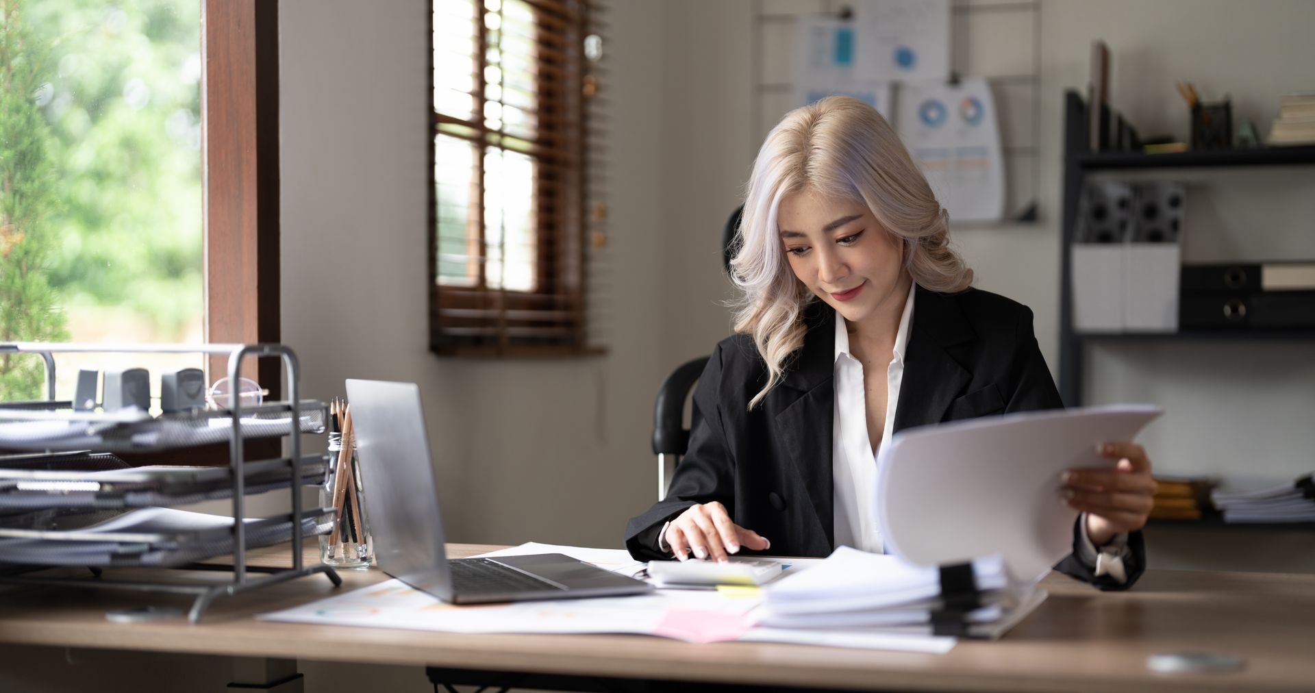Person working at a desk in a bright office, reviewing papers beside a laptop and printer.