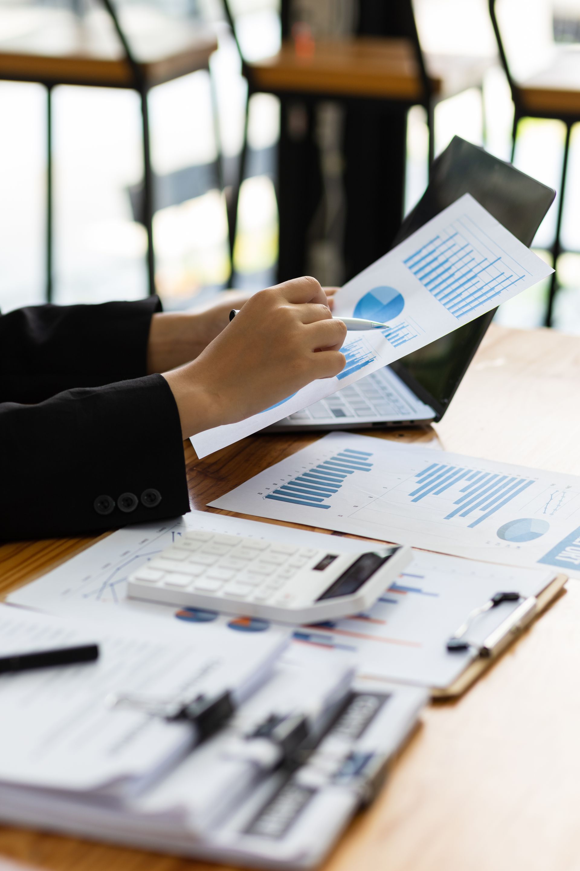 Hand holding a tablet over printed charts and a calculator on a desk in an office setting
