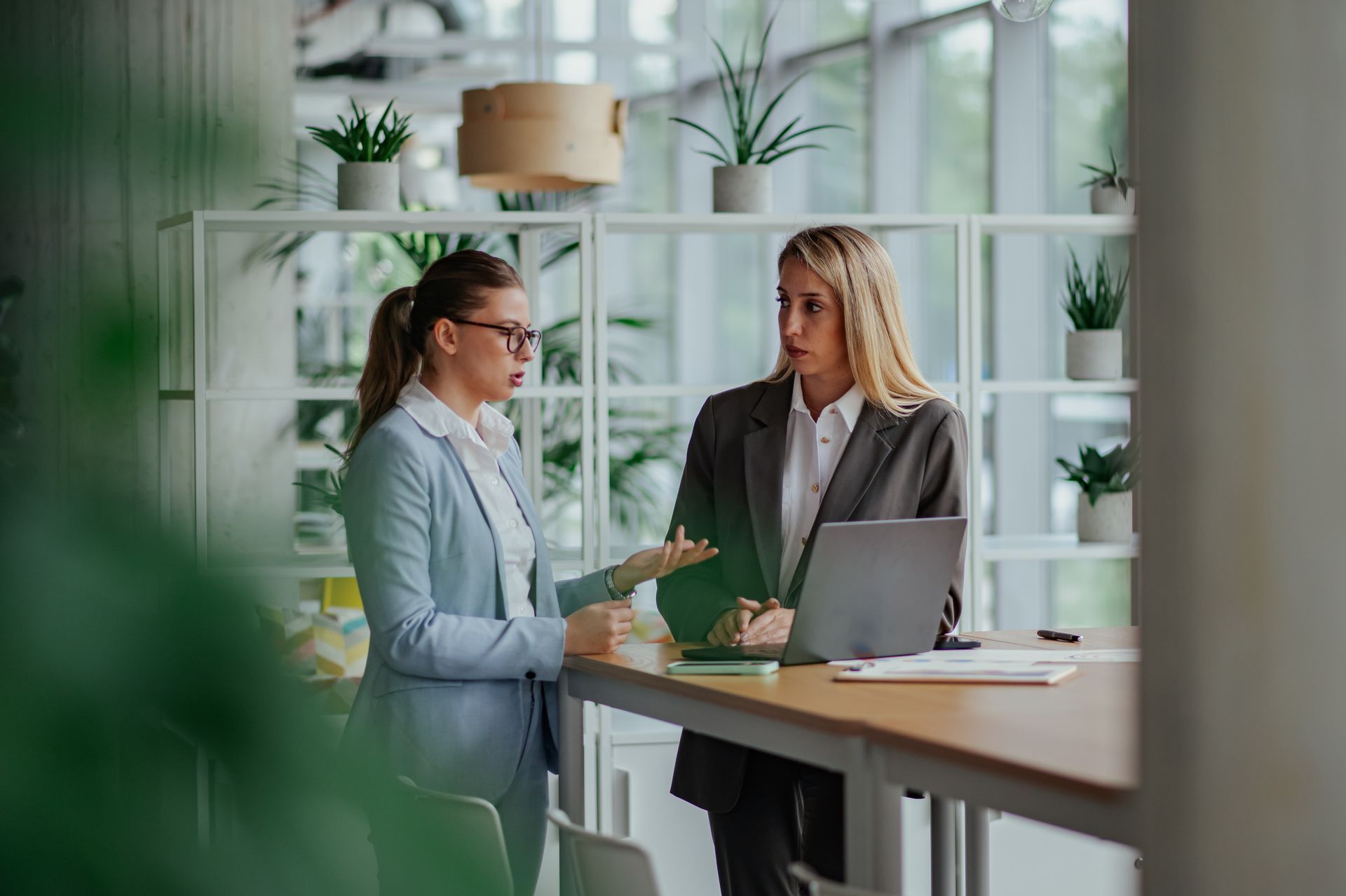 Two professionals talk at a bright office counter, one gesturing beside a laptop and papers.