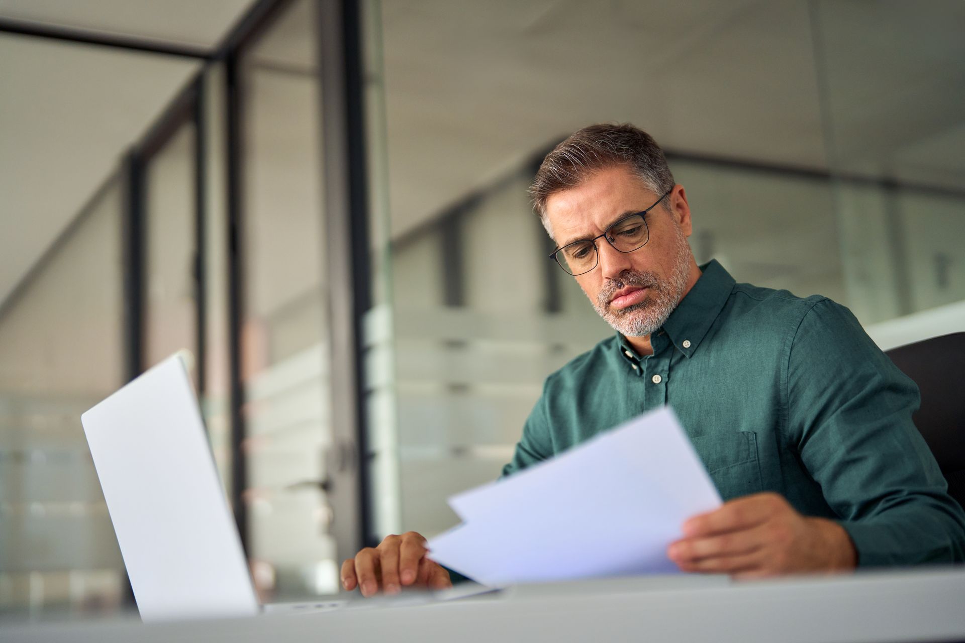 Man at a desk reviewing papers beside an open laptop in a modern office