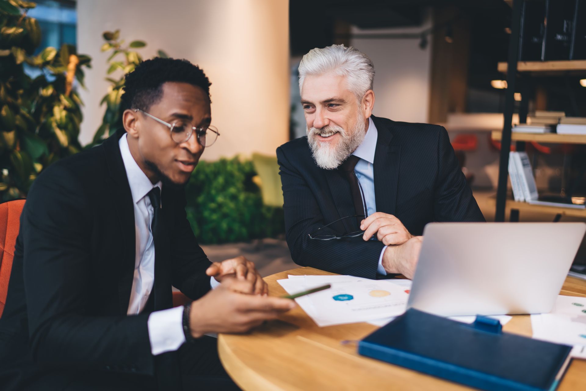 Two professionals in suits sitting at a round table, discussing documents and looking at a smartphone in an office.
