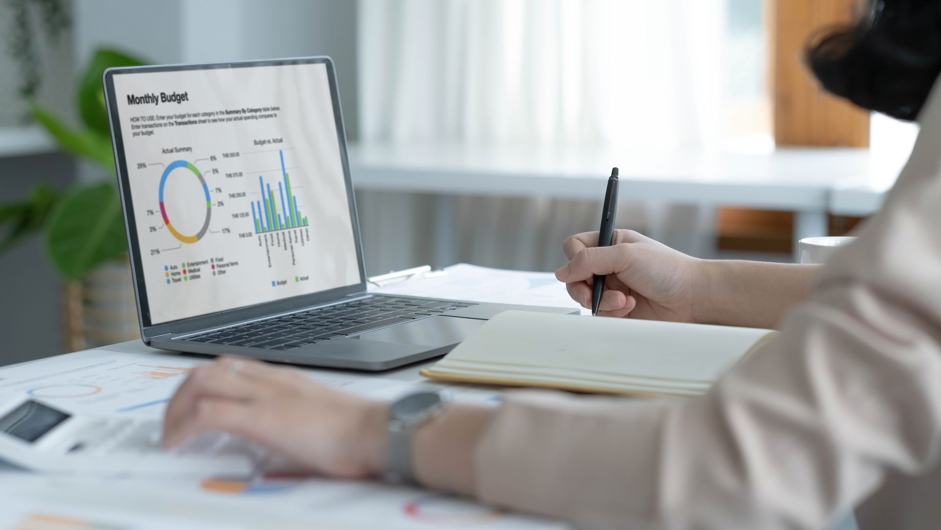 Person analyzing charts on a laptop while taking notes at a desk in a bright office